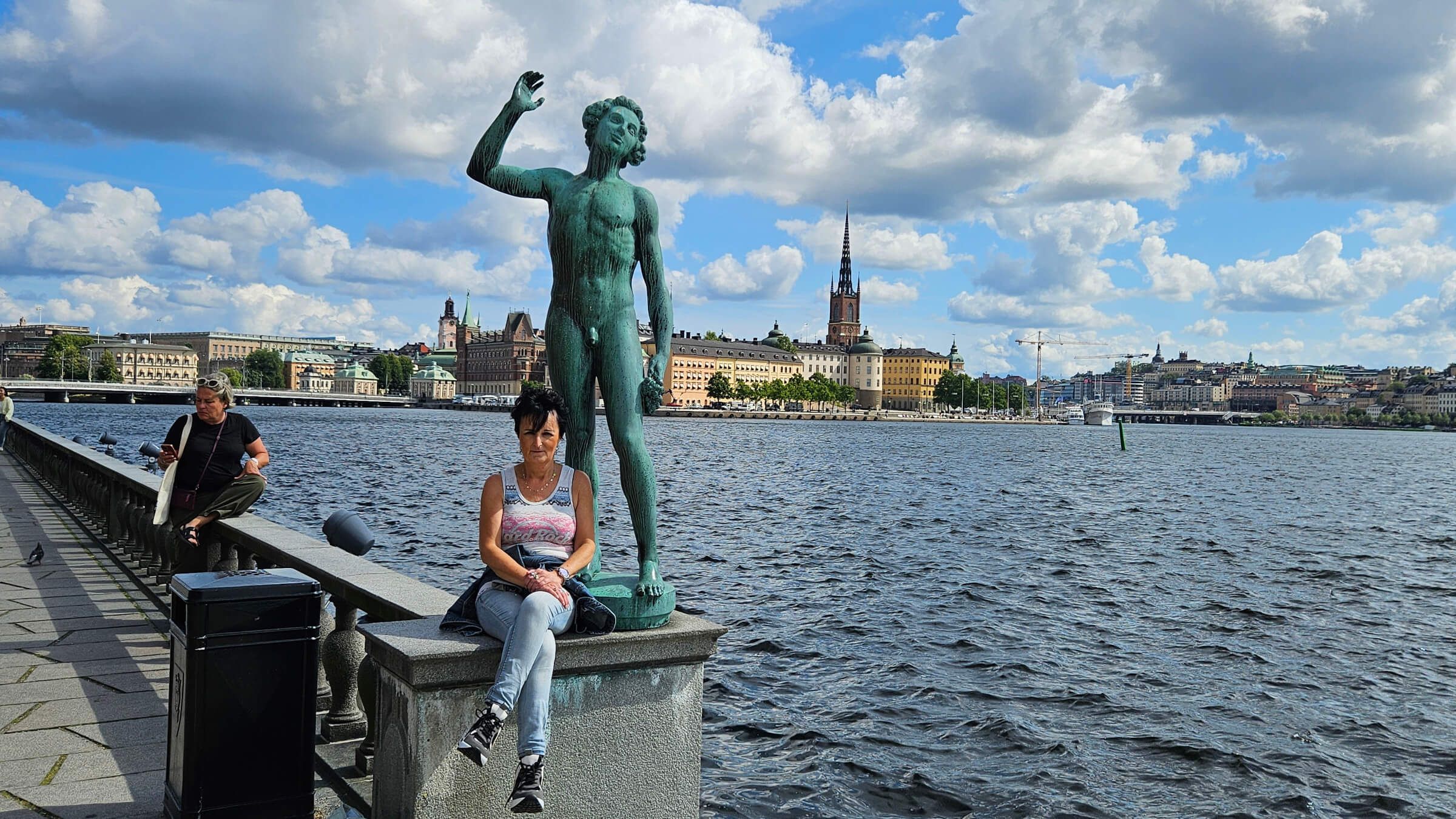 Eine Statue am Wasser mit einer Frau, die sitzt, während eine Stadtlandschaft im Hintergrund sichtbar ist.
