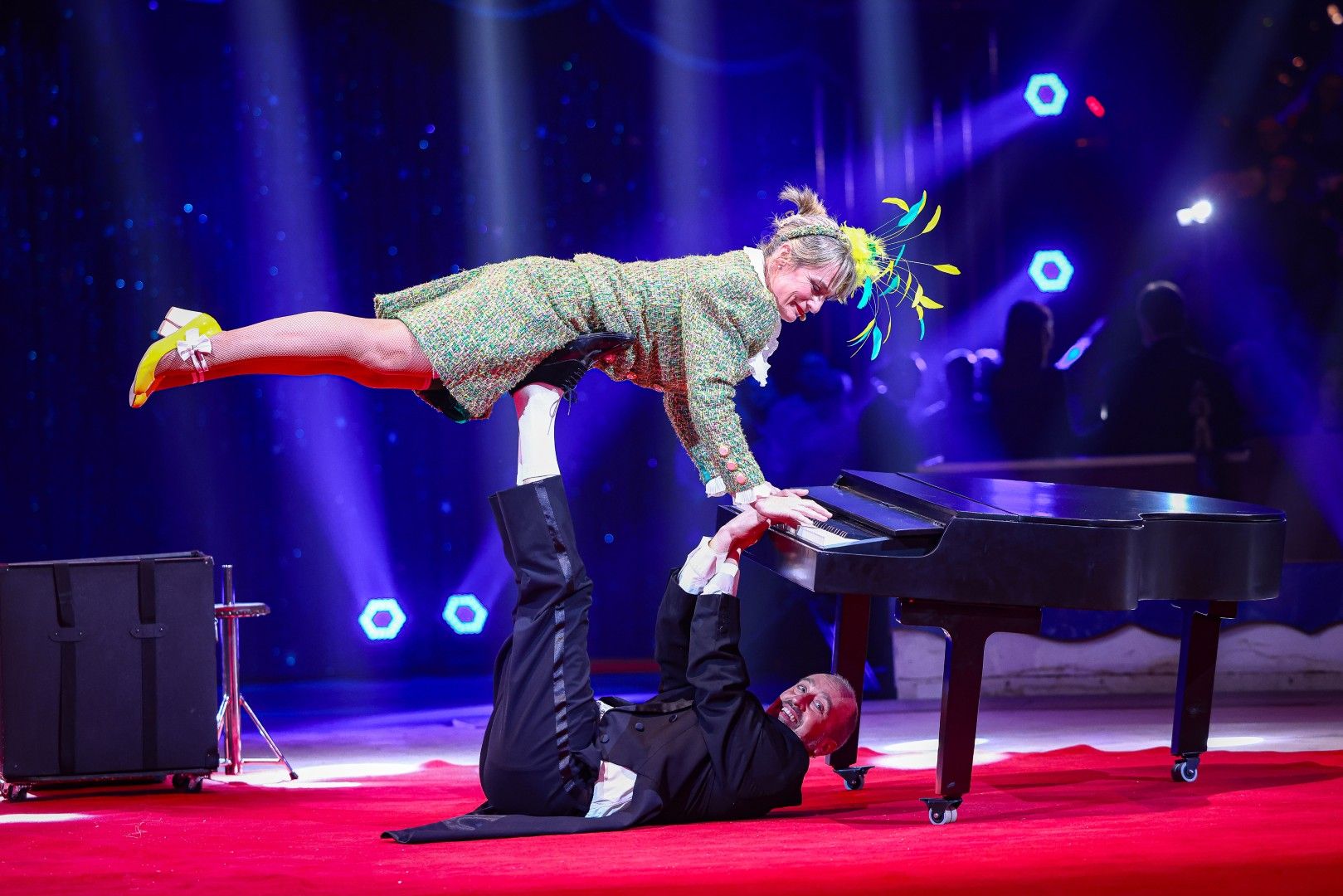 Ein Clown spielt Klavier, während eine farbenfroh gekleidete Akrobatin darauf balanciert. Zirkusatmosphäre.