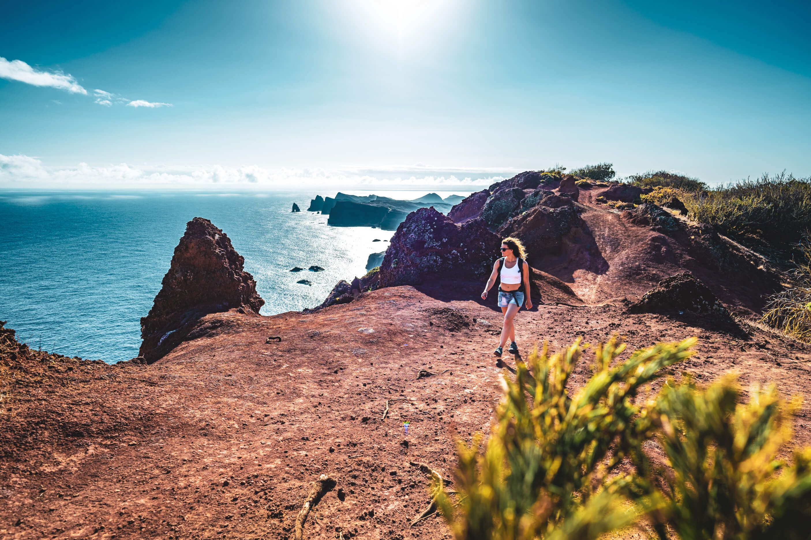 Eine Frau wandert auf einem felsigen Weg mit Blick auf das glitzernde Meer und die Küste im Hintergrund.