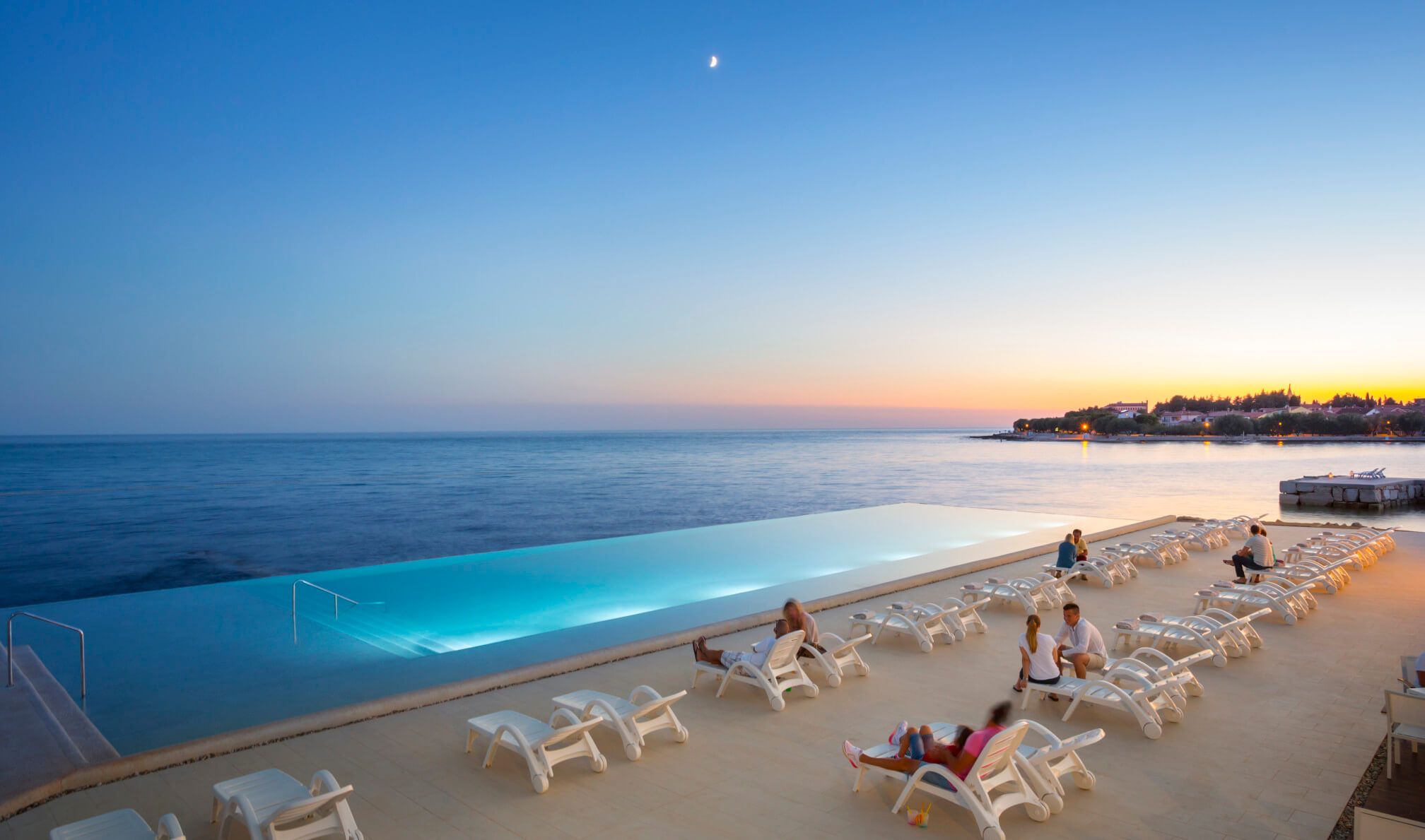 Ein Pool mit Blick auf das Meer bei Sonnenuntergang, Sonnenliegen und klare Himmel mit Mond. Entspannende Atmosphäre.