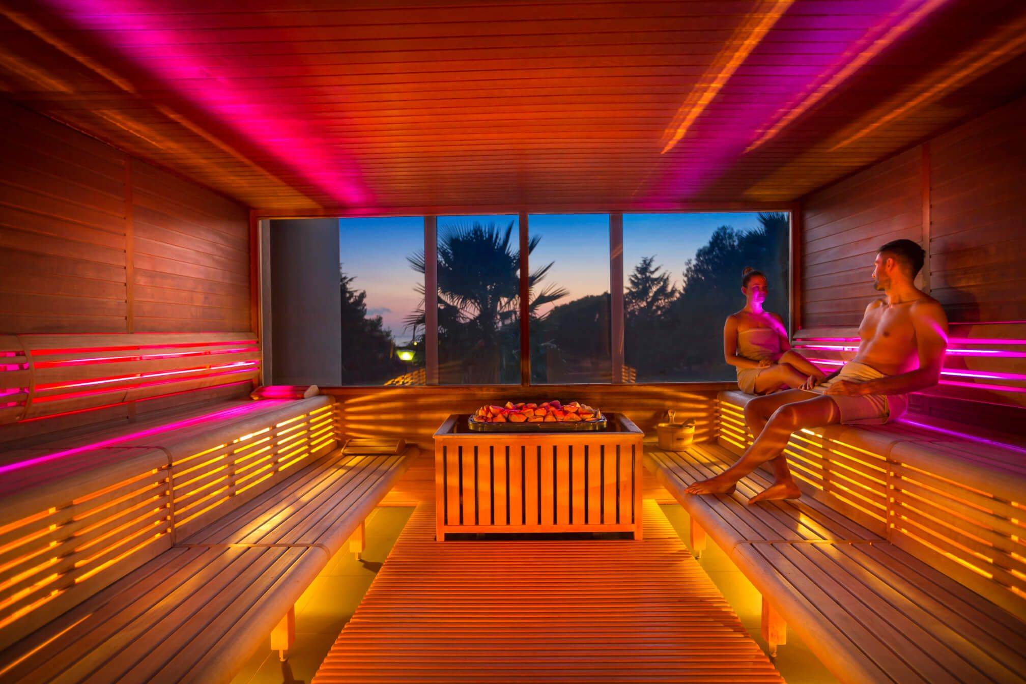 Zwei Personen entspannen in einer modernen Sauna mit farbigem Licht und Blick auf den Sonnenuntergang.