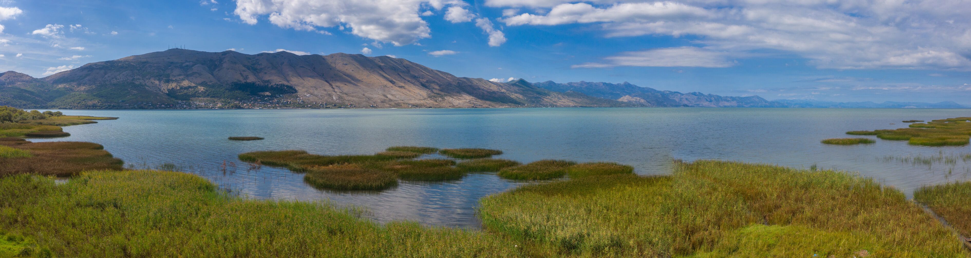 Ein malerischer See umgeben von Bergen, mit grünem Schilf und blauem Himmel, ideal für Naturliebhaber.