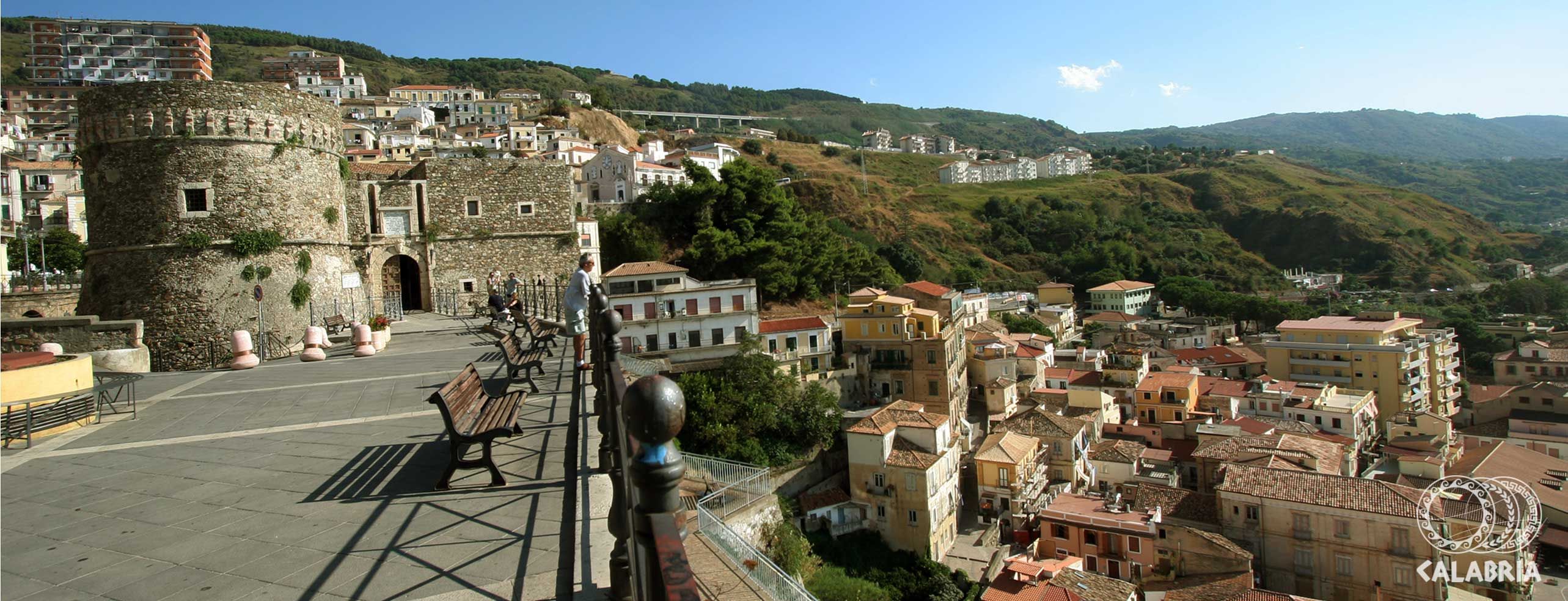 Eine Panoramaansicht eines historischen Ortes mit Steinturm und farbenfrohen Gebäuden unter blauem Himmel.