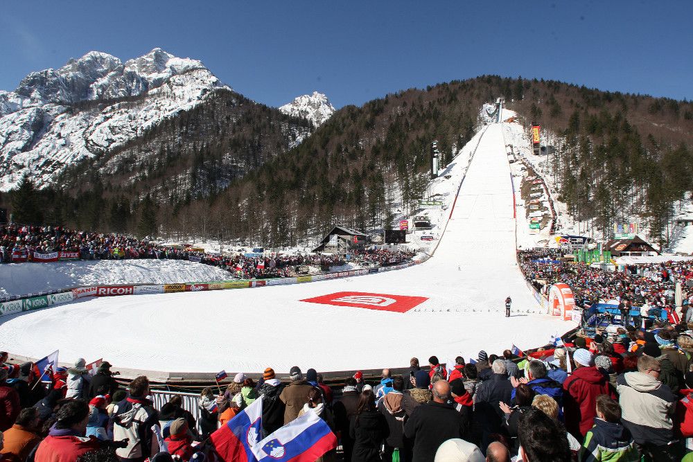 Ein Skisprungareal mit Zuschauern, schneebedeckten Bergen und blauen Himmel. Aufregende Wettkampfstimmung!