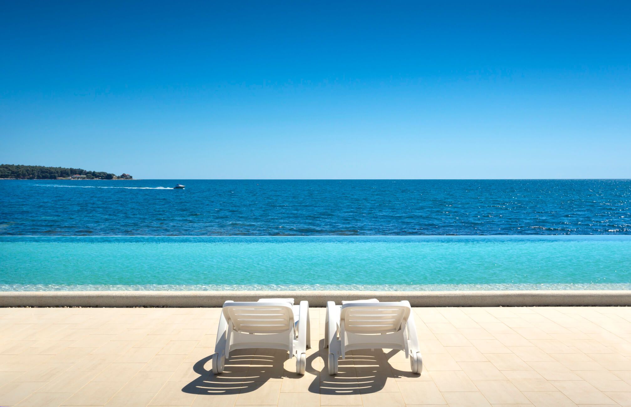 Zwei Liegen stehen am Wasser, mit Blick auf ein weites, blaues Meer unter klarem Himmel. Ein Boot fährt vorbei.
