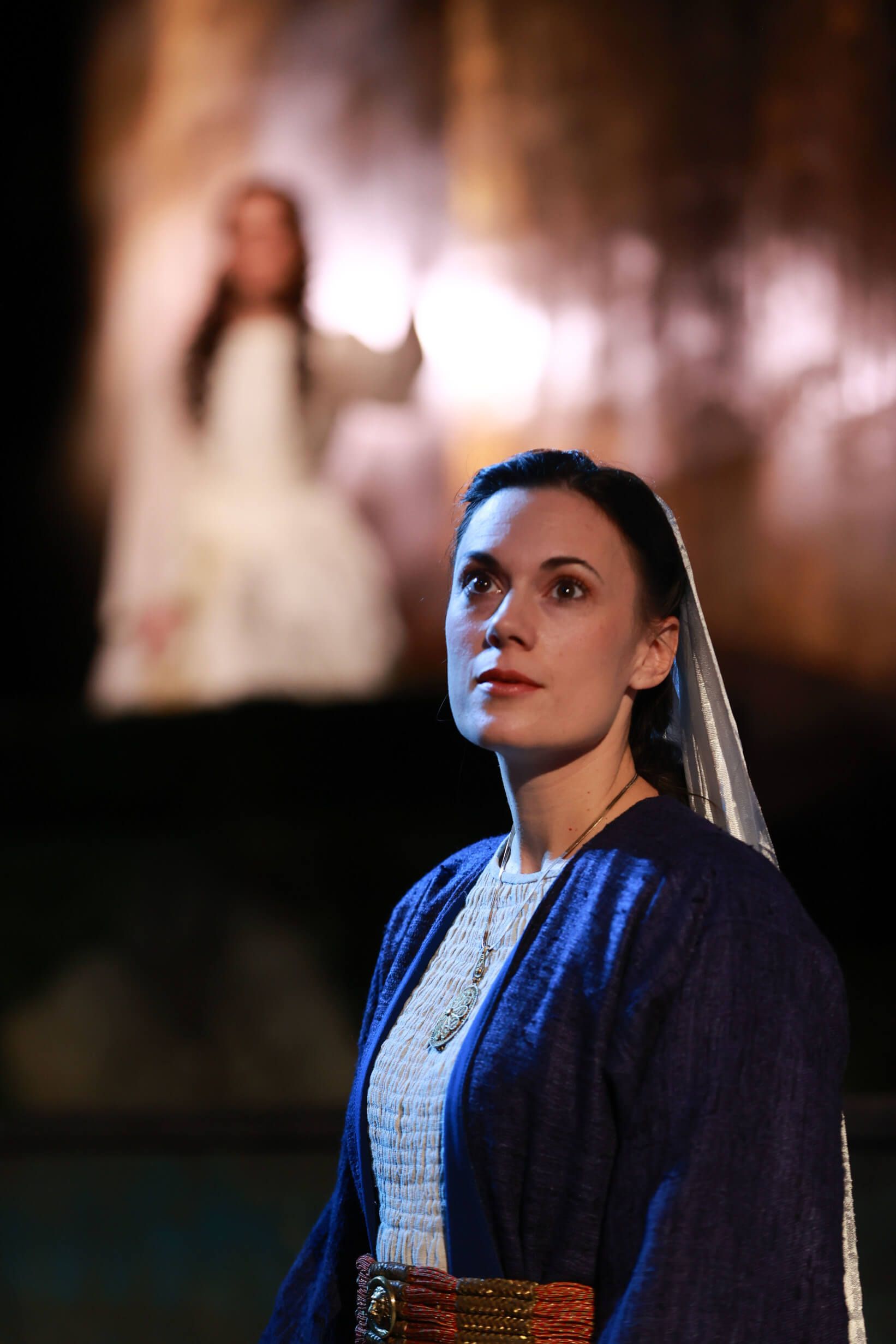 Eine Frau in historischem Gewand schaut gespannt nach oben, während eine andere Figur im Hintergrund zu sehen ist.