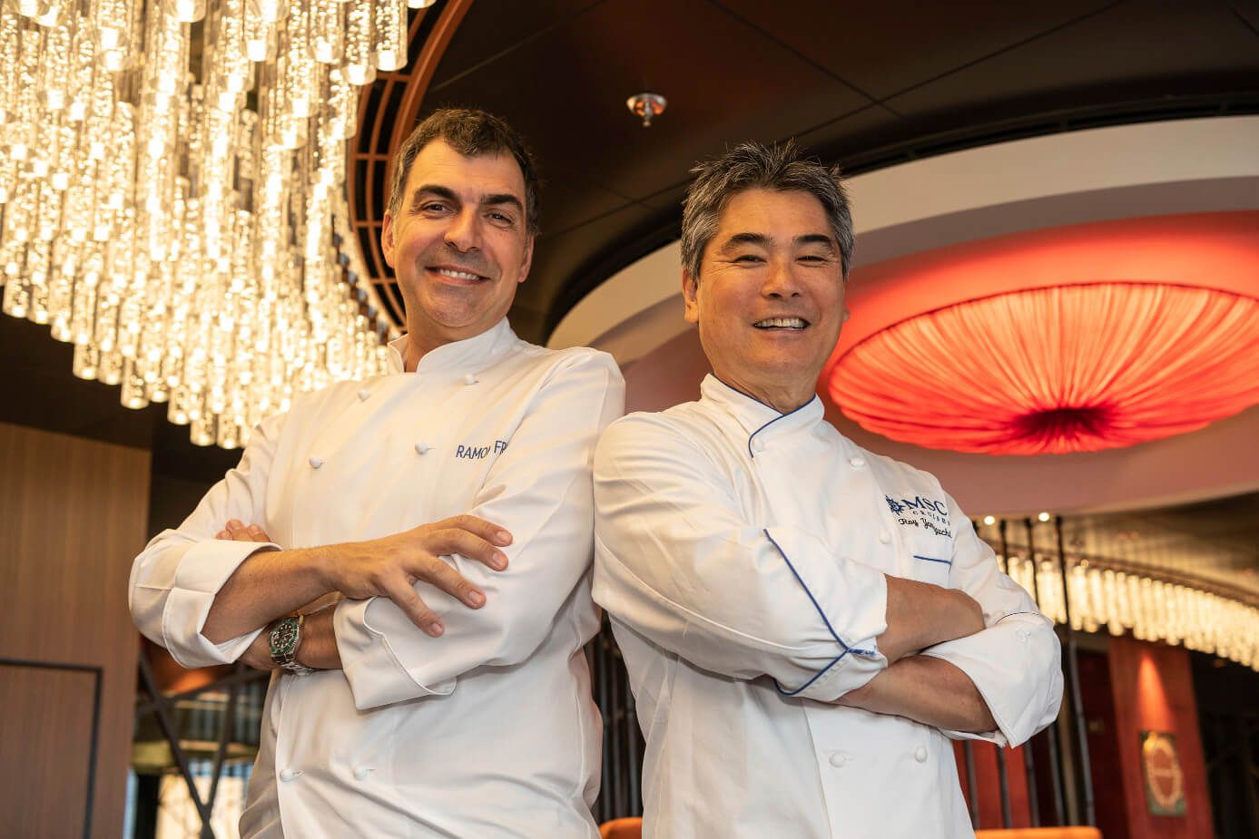 Zwei Köche lächeln in einer eleganten Umgebung unter einer beeindruckenden Deckenleuchte.