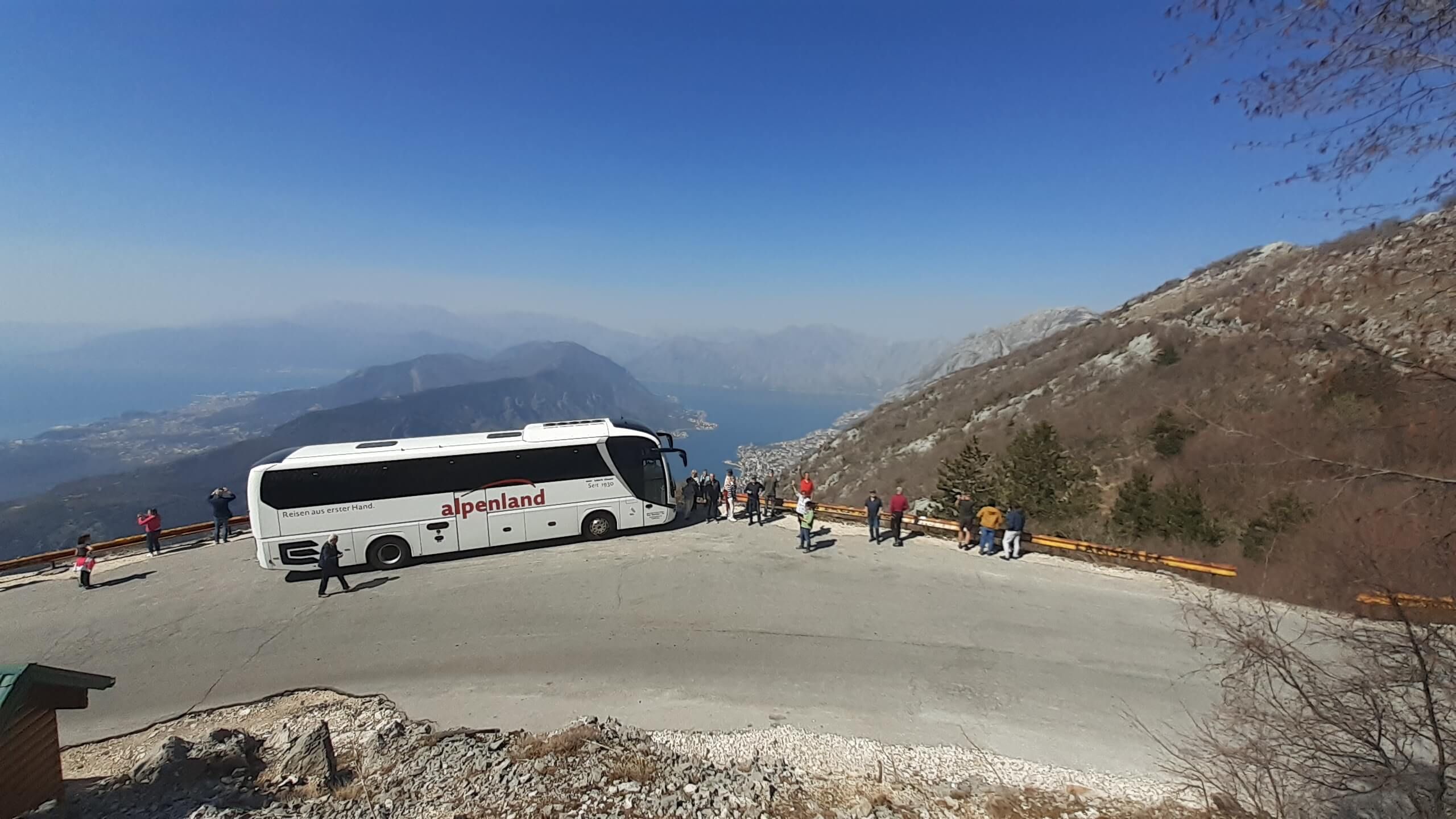 Ein Reisebus steht an einem Aussichtspunkt mit Blick auf Berge und einen See, viele Touristen machen Fotos.