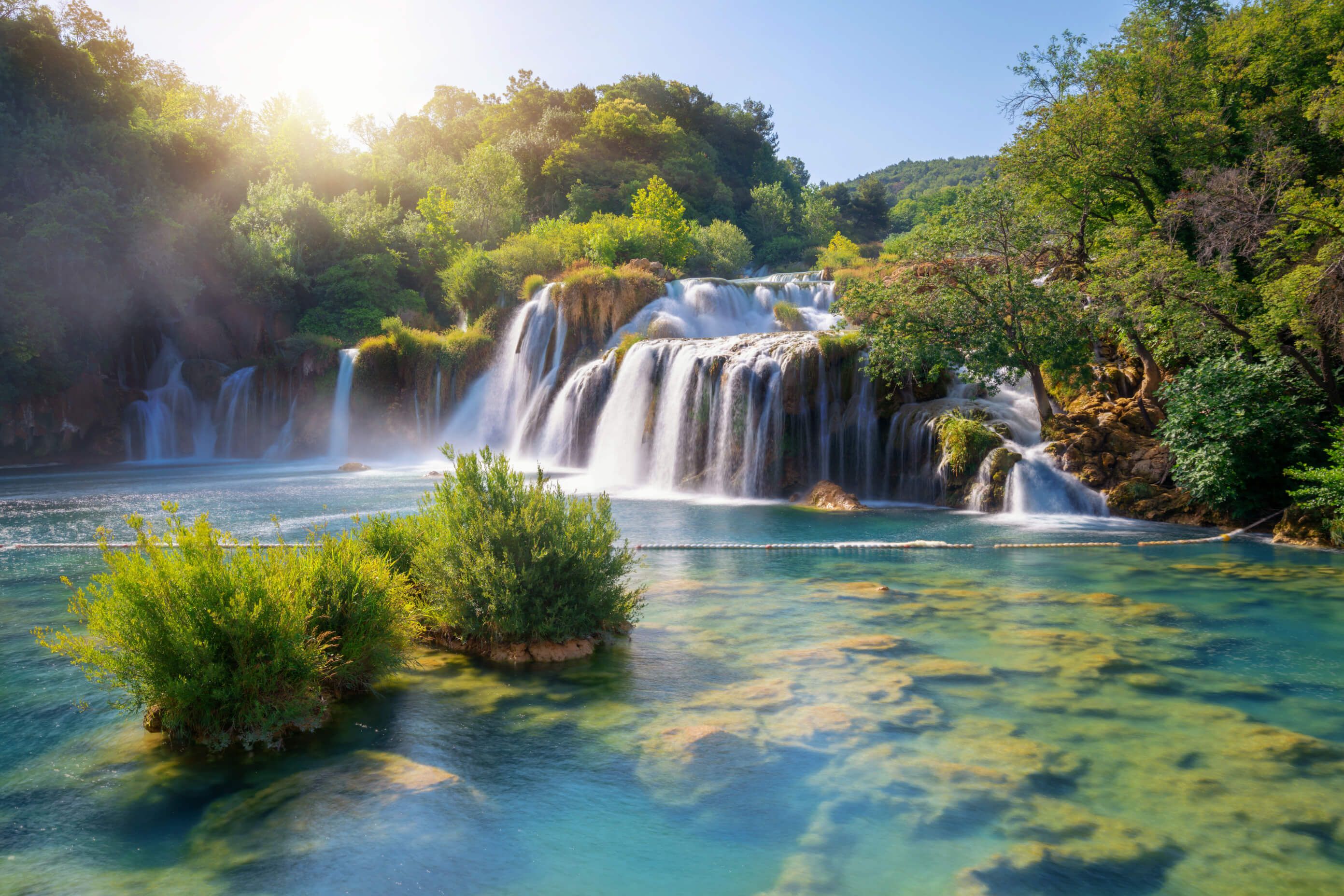 Ein malerischer Wasserfall im Nationalpark Plitvice sprudelt in ruhigen, klaren Gewässern, umgeben von üppigem Grün und Sonnenlicht.