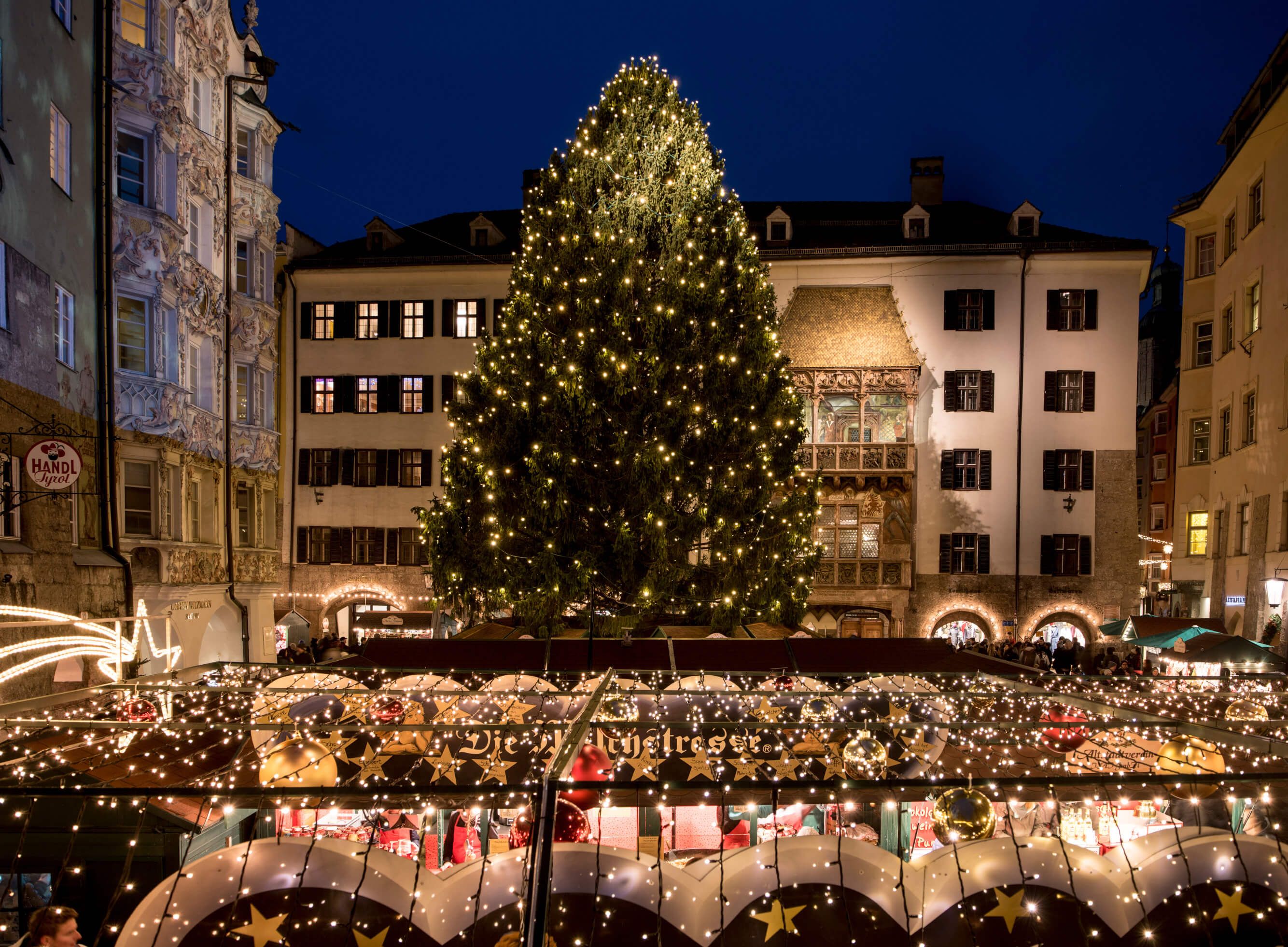 Ein festlich geschmückter Weihnachtsmarkt mit einem großen, beleuchteten Weihnachtsbaum bei Nacht.
