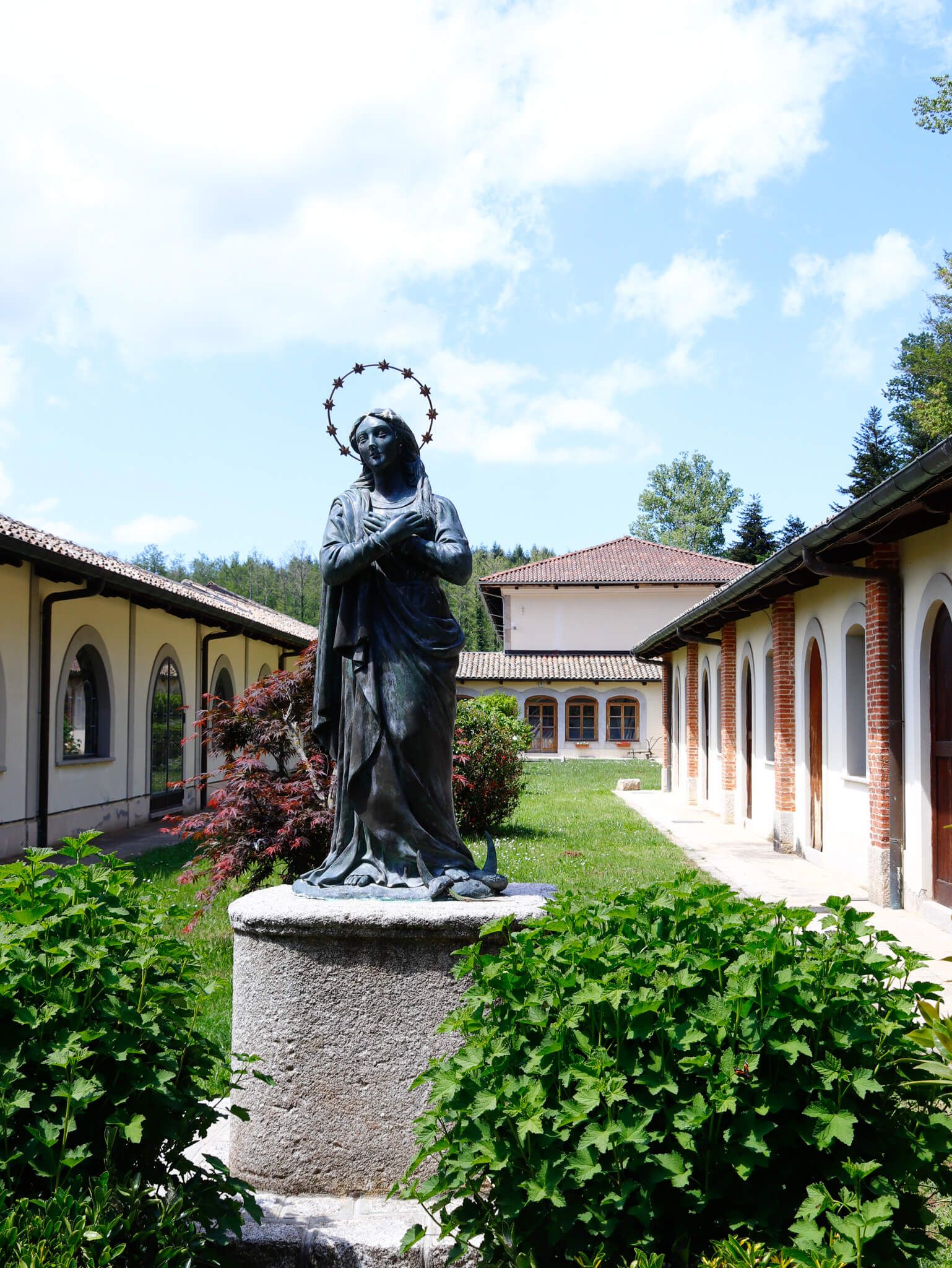 Eine Statue einer heiligen Figur steht in einem ruhigen Garten umgeben von Gebäuden und grünem Gras.