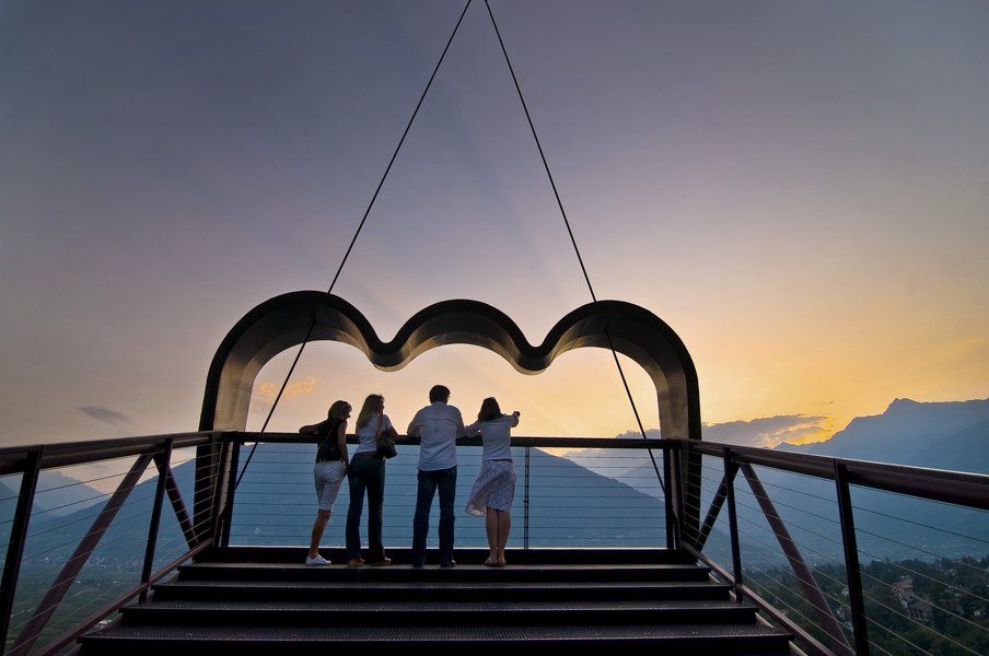 Vier Personen stehen auf einer Aussichtsplattform und schauen auf eine malerische Berglandschaft bei Sonnenuntergang.