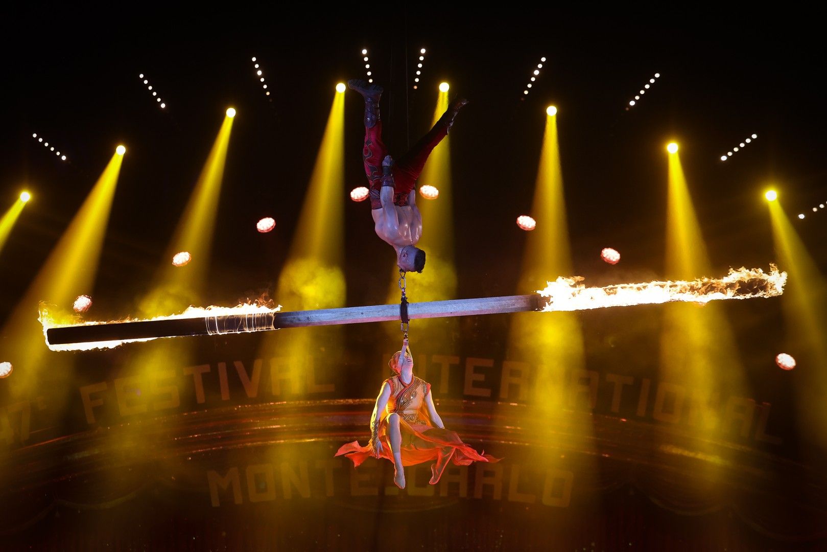 Akrobaten schweben an einem feurigen Balken, umgeben von bunten Lichtern in einer beeindruckenden Show.