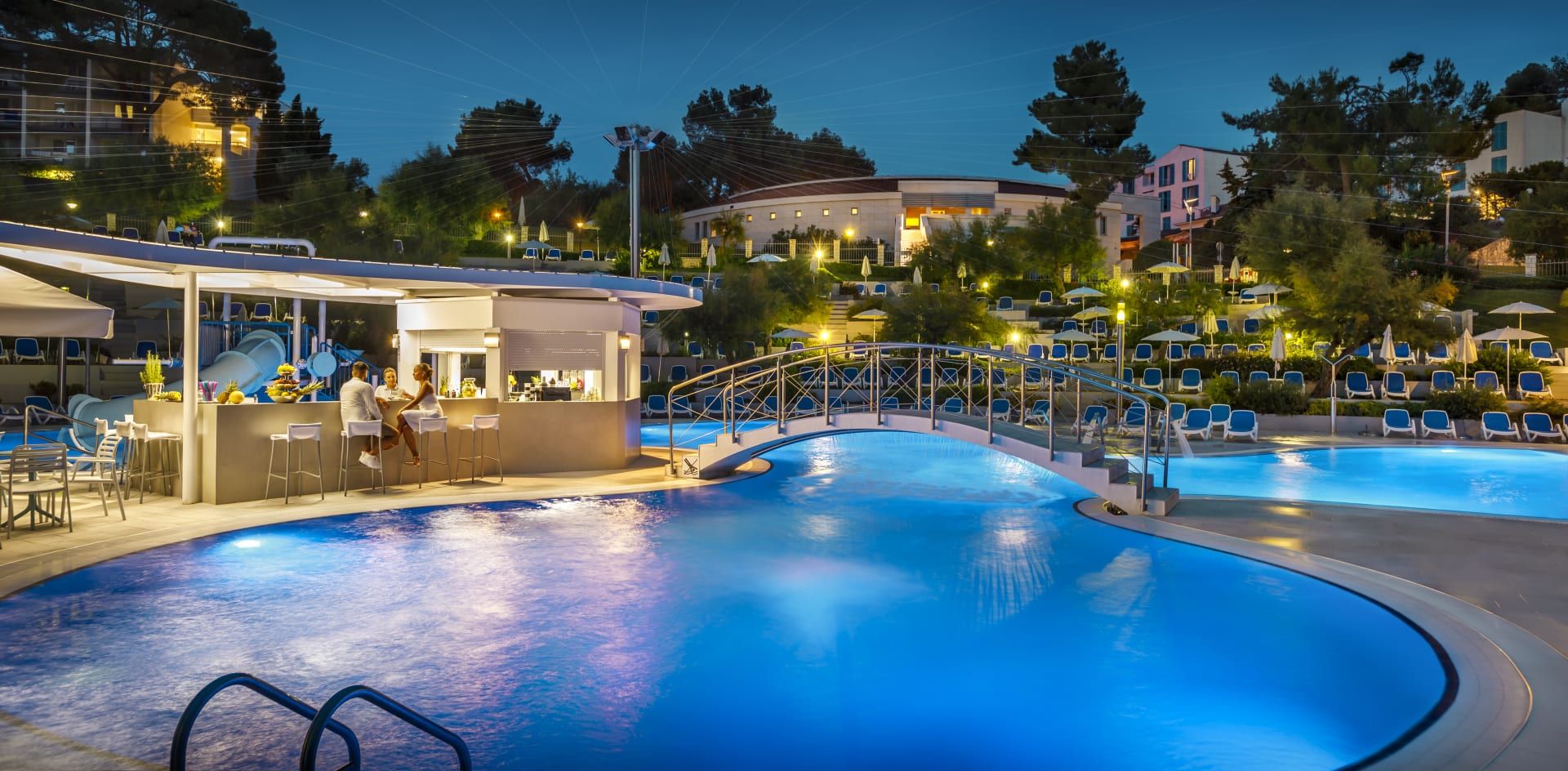 Ein beleuchteter Poolbereich mit Bar, Liegen und einer Brücke bei Nacht, umgeben von Bäumen und Gebäuden.