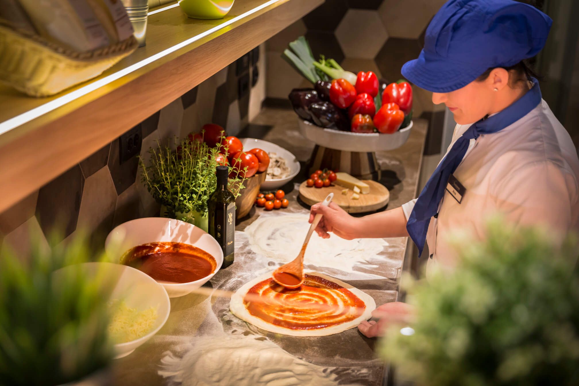 Eine Köchin bereitet einen Pizzaboden vor und verteilt Tomatensauce in einer modernen Küche. Frisches Gemüse ist sichtbar.