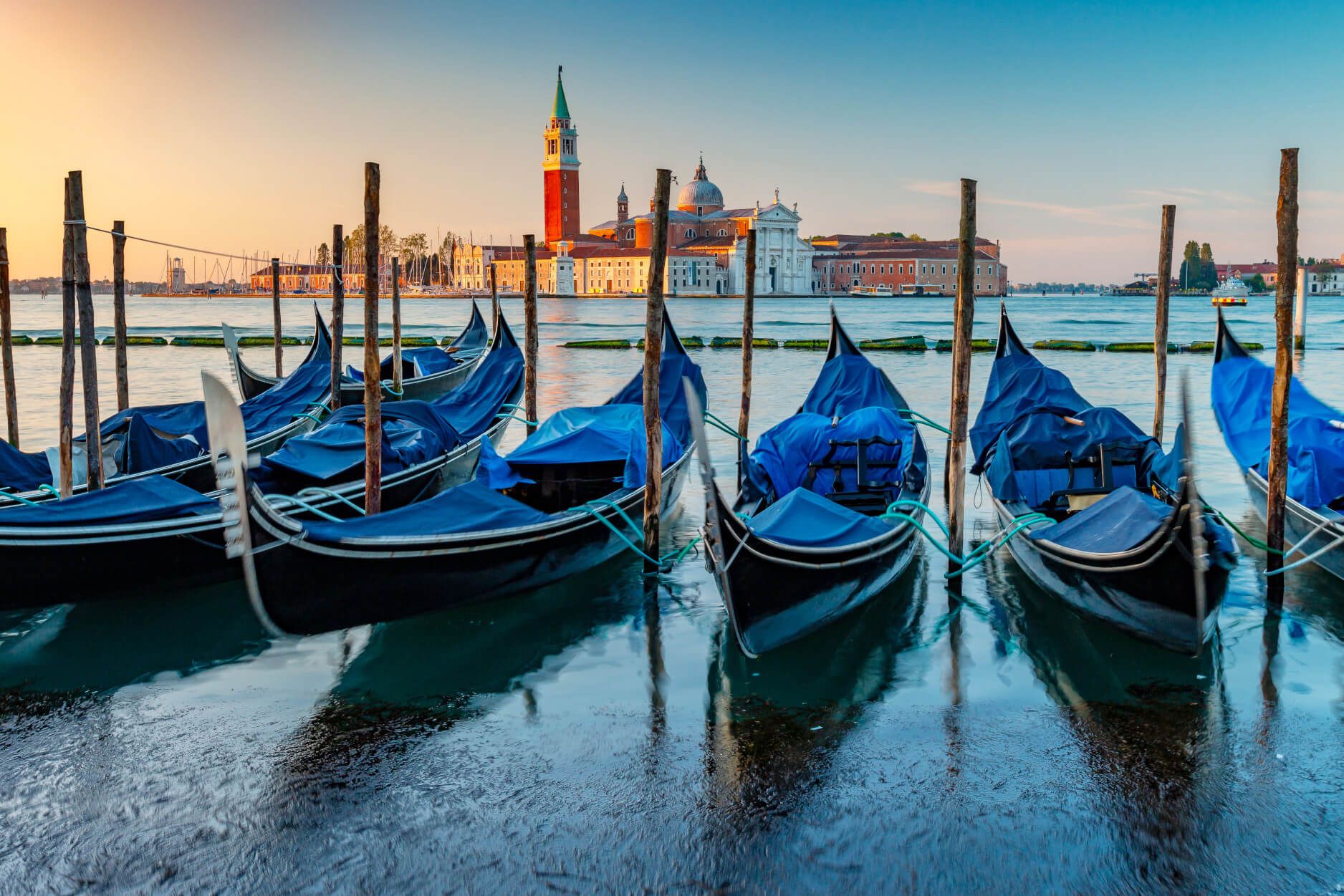 Gondeln mit blauen Abdeckungen am Wasser, im Hintergrund die Architektur Venetiens zum Sonnenaufgang.