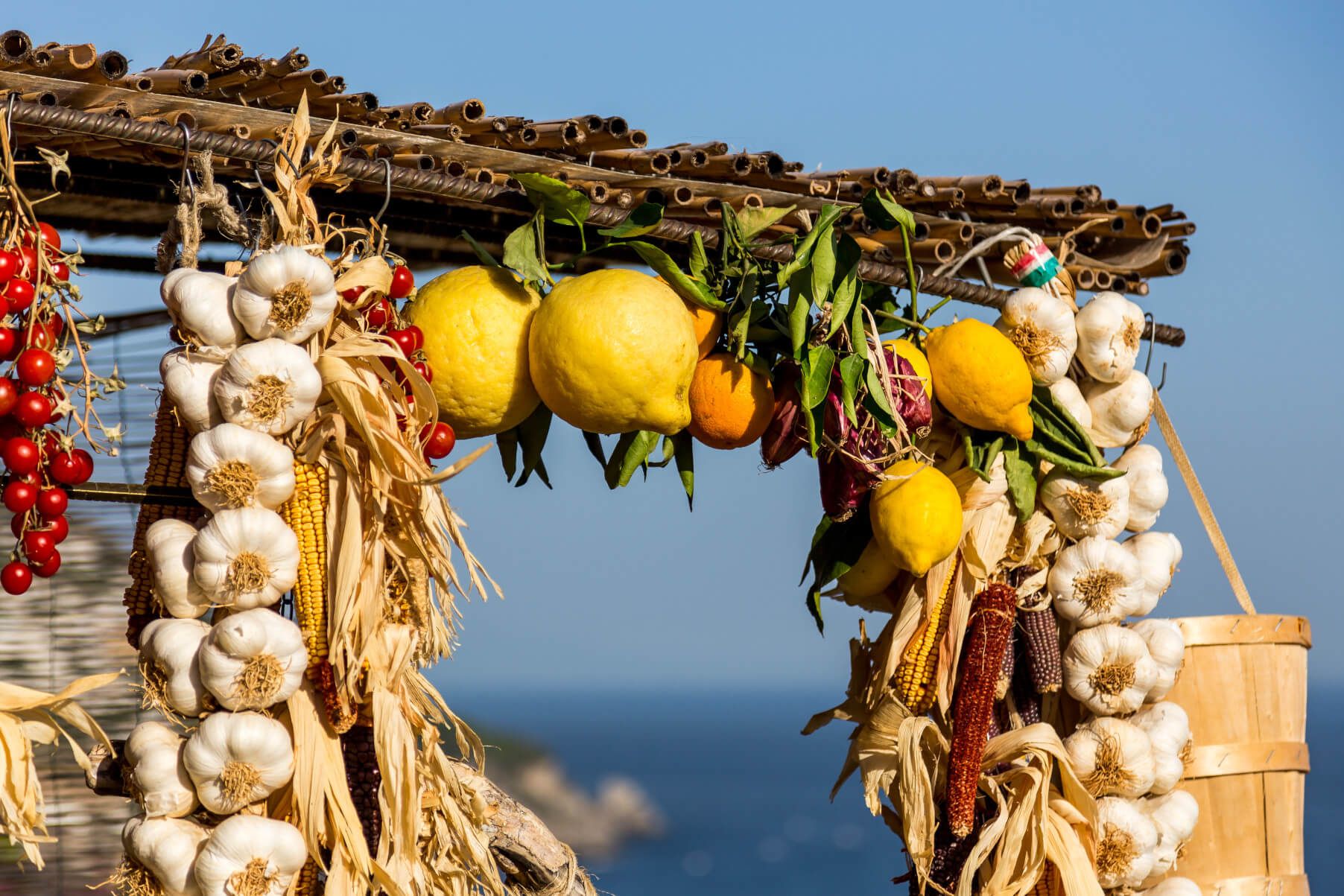 Bunte Früchte und Knoblauch hängen dekorativ unter einem Dach, mit blauen Himmel und Meer im Hintergrund.