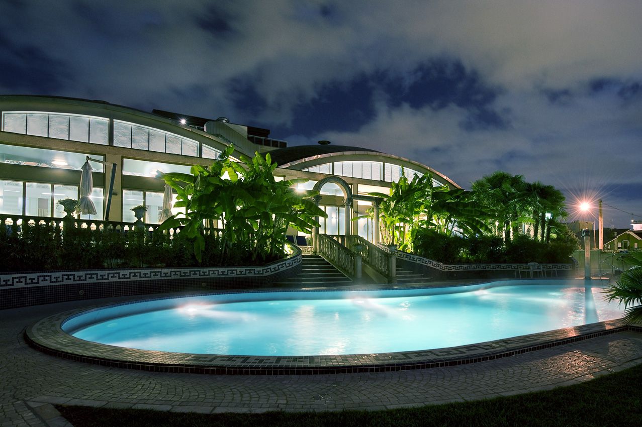 Ein beleuchteter Pool umgeben von tropischen Pflanzen und einem modernen Gebäude bei Nacht.