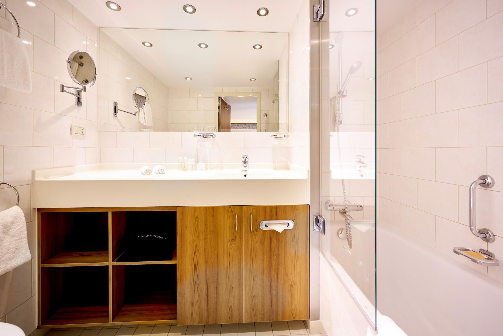 Ein modernes Badezimmer mit Holzregal, Waschbecken und klaren Kanten. Helle Fliesen und große Spiegel sorgen für Weite.