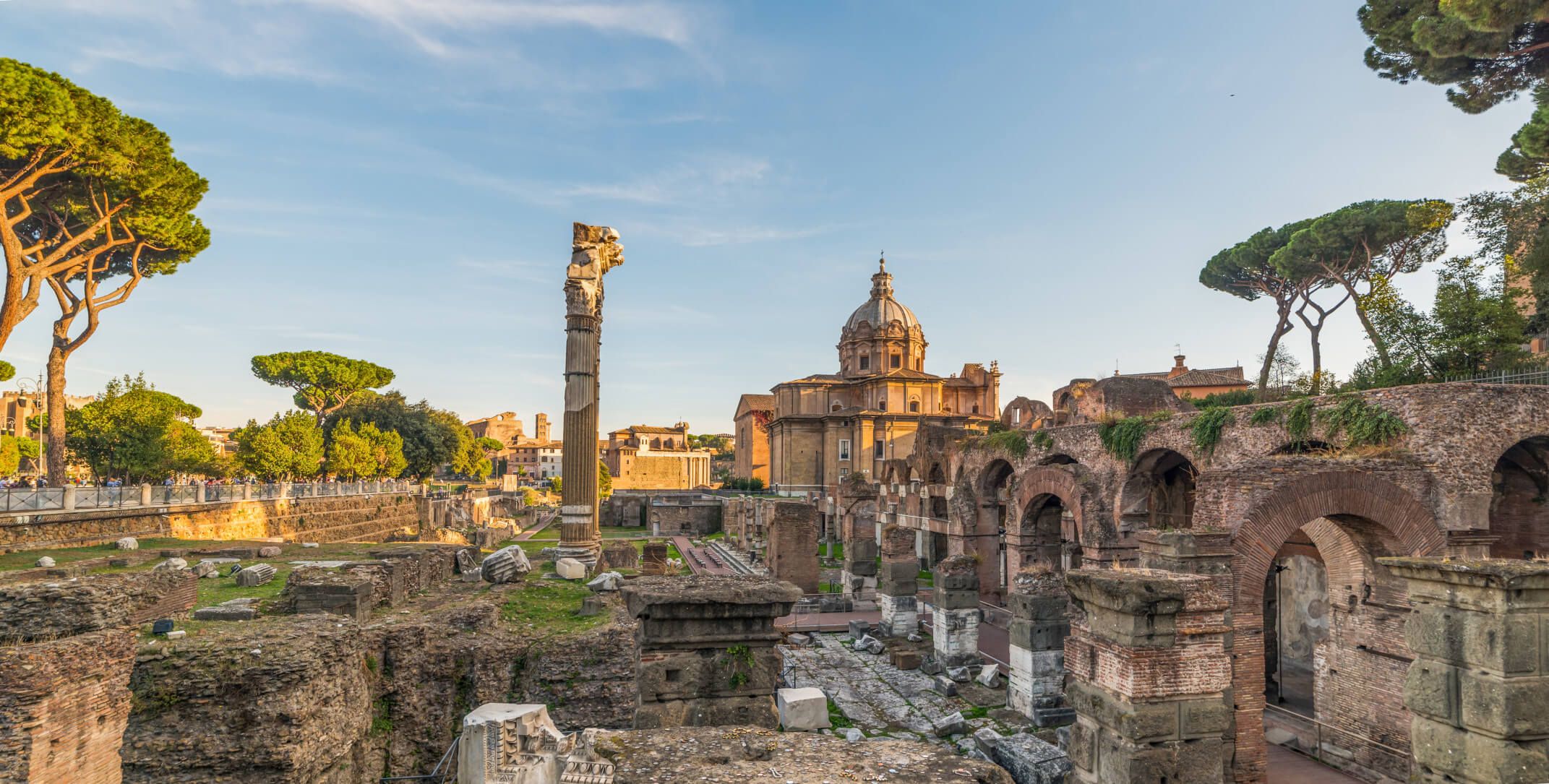 Römische Ruinen mit einem Säulenfragment, Bäumen und historischen Gebäuden unter blauem Himmel.