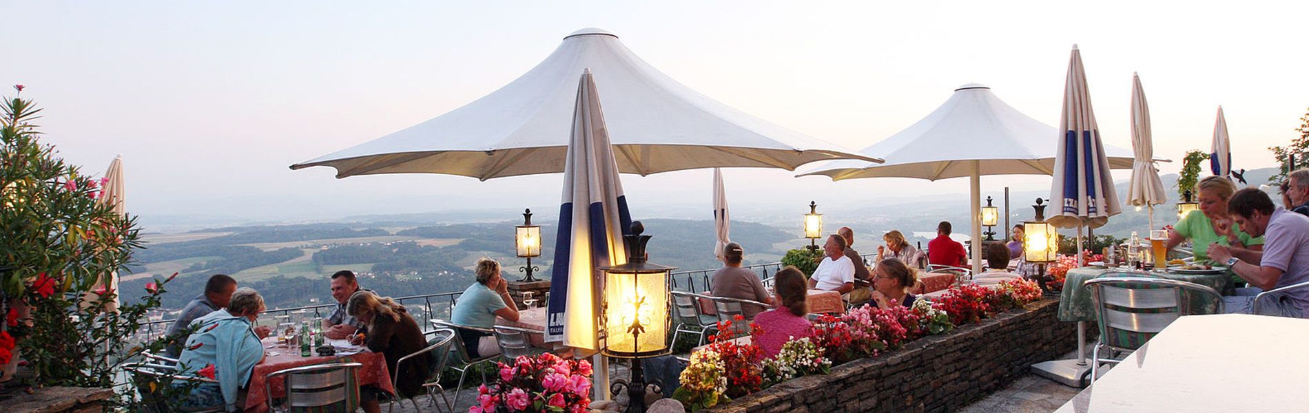 Gemütliche Terrasse mit Blick ins Tal, Gäste genießen Speisen und Getränke unter Sonnenschirmen bei Dämmerung.