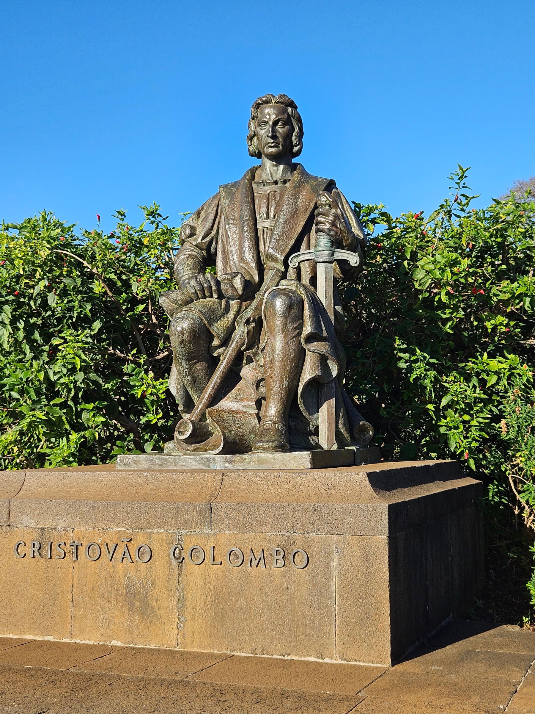 Eine Statue von Cristóvão Colombo sitzt auf einem Podest, umgeben von grünen Sträuchern und blauem Himmel.