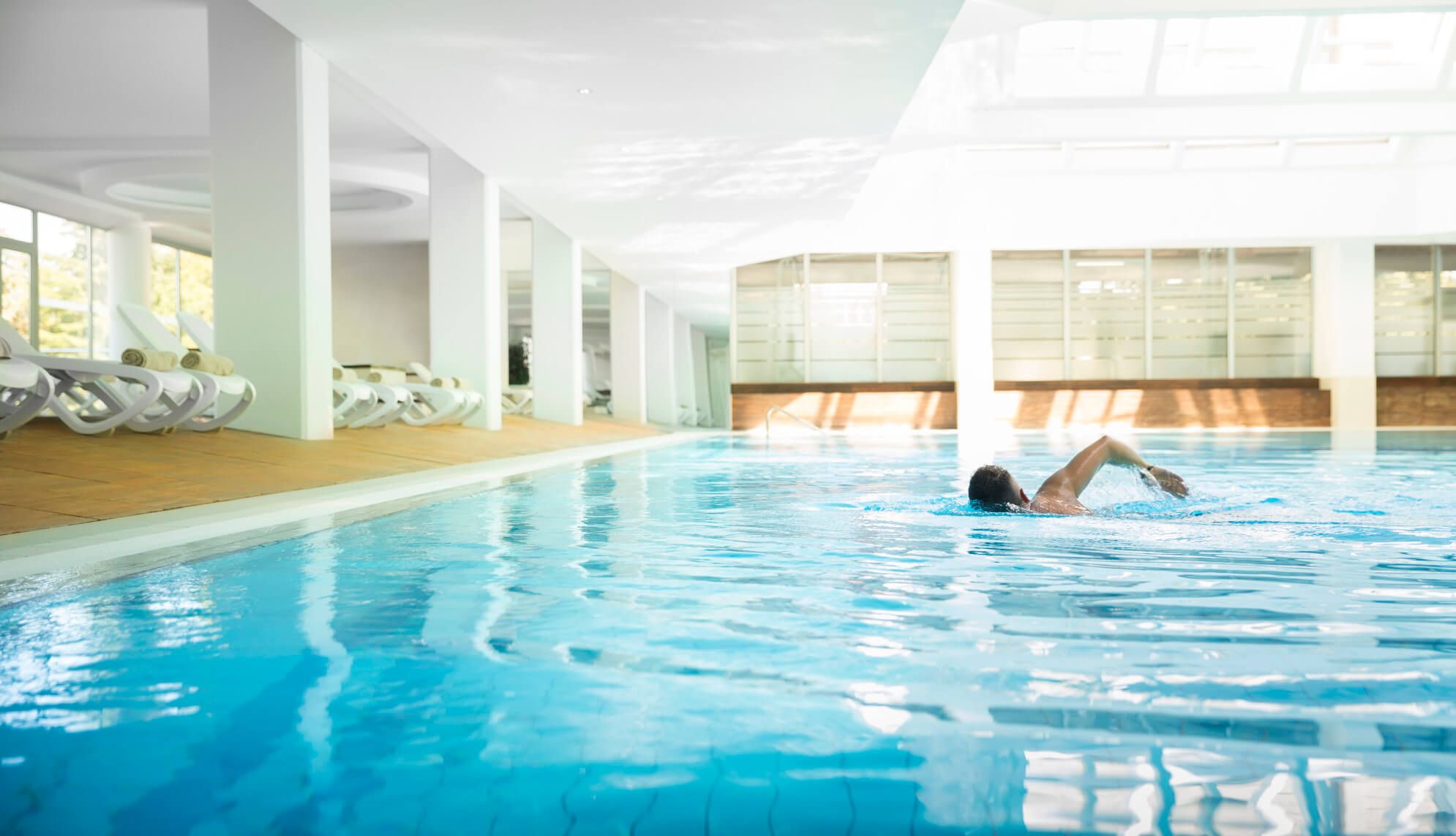 Ein Hallenbad mit klarem Wasser. Eine Person schwimmt ruhig, Sonnenlicht fällt durch große Fenster. Entspannende Atmosphäre.