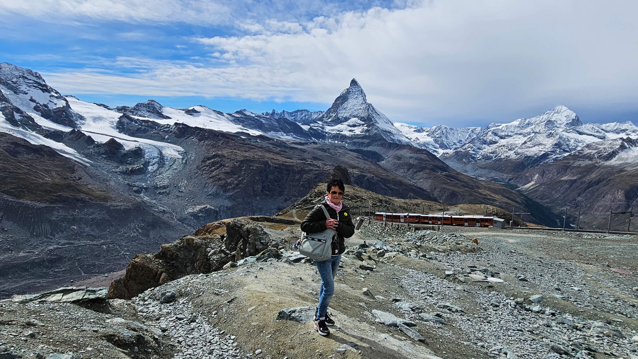 Eine Person steht auf einem steinigen Weg mit dem Matterhorn und schneebedeckten Bergen im Hintergrund.