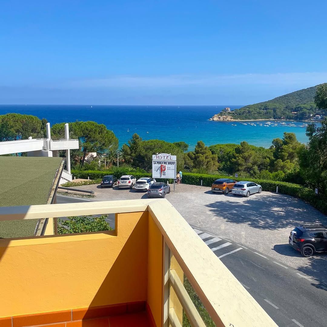 Blick von einem Balkon auf das blaue Meer, Strände und eine ruhige Parklandschaft mit mehreren Autos.