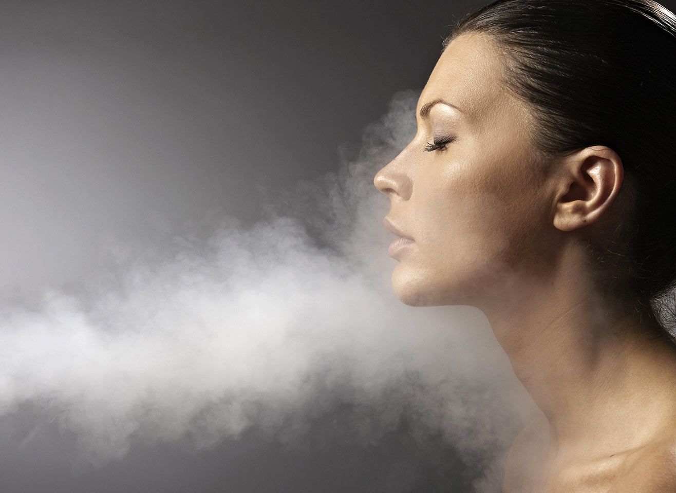 Eine Frau mit geschlossenen Augen, aus deren Mund Rauch strömt, vor einem grauen Hintergrund.