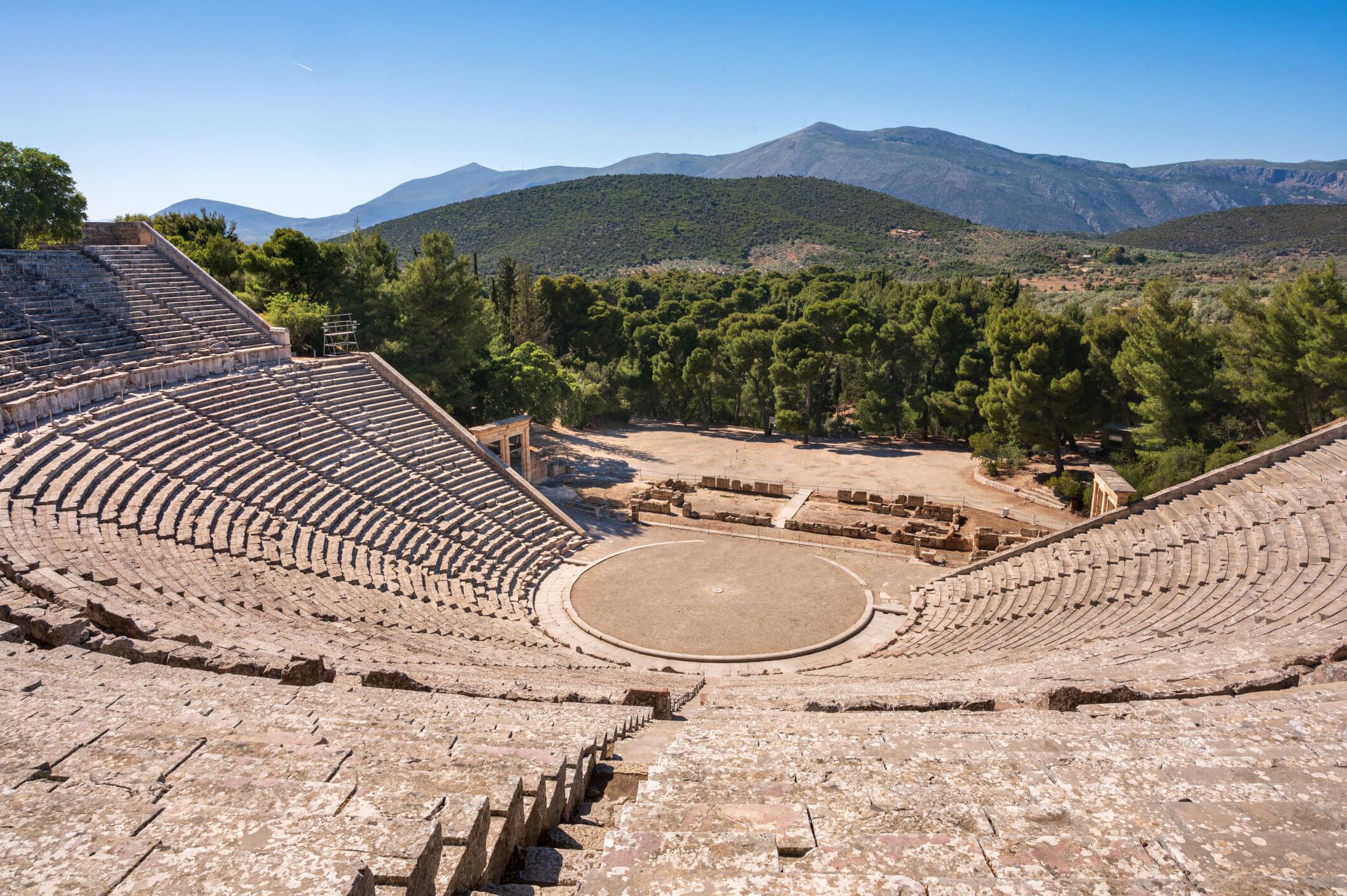 Ein antikes Theater in einer grünen Landschaft, umgeben von sanften Hügeln und blauem Himmel.