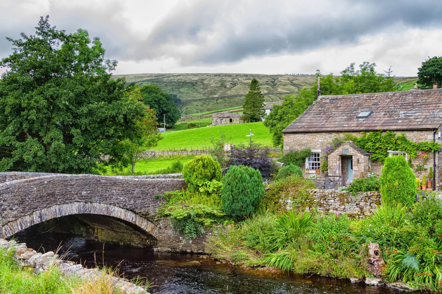 Ein ruhiges, ländliches Bild mit einem Steinhus, Brücke und grünen Wiesen unter einem bewölkten Himmel.