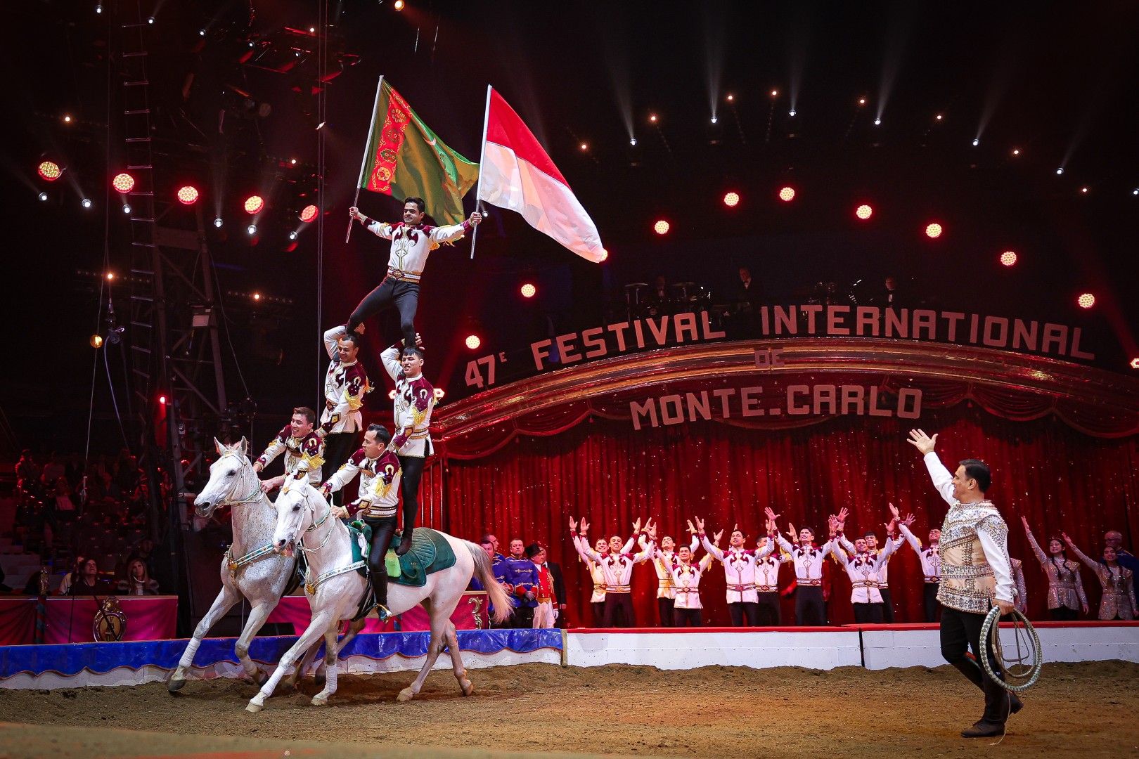 Ein Zirkusauftritt mit Reitern, die Fahnen schwenken, auf weißen Pferden, umgeben von jubelndem Publikum.