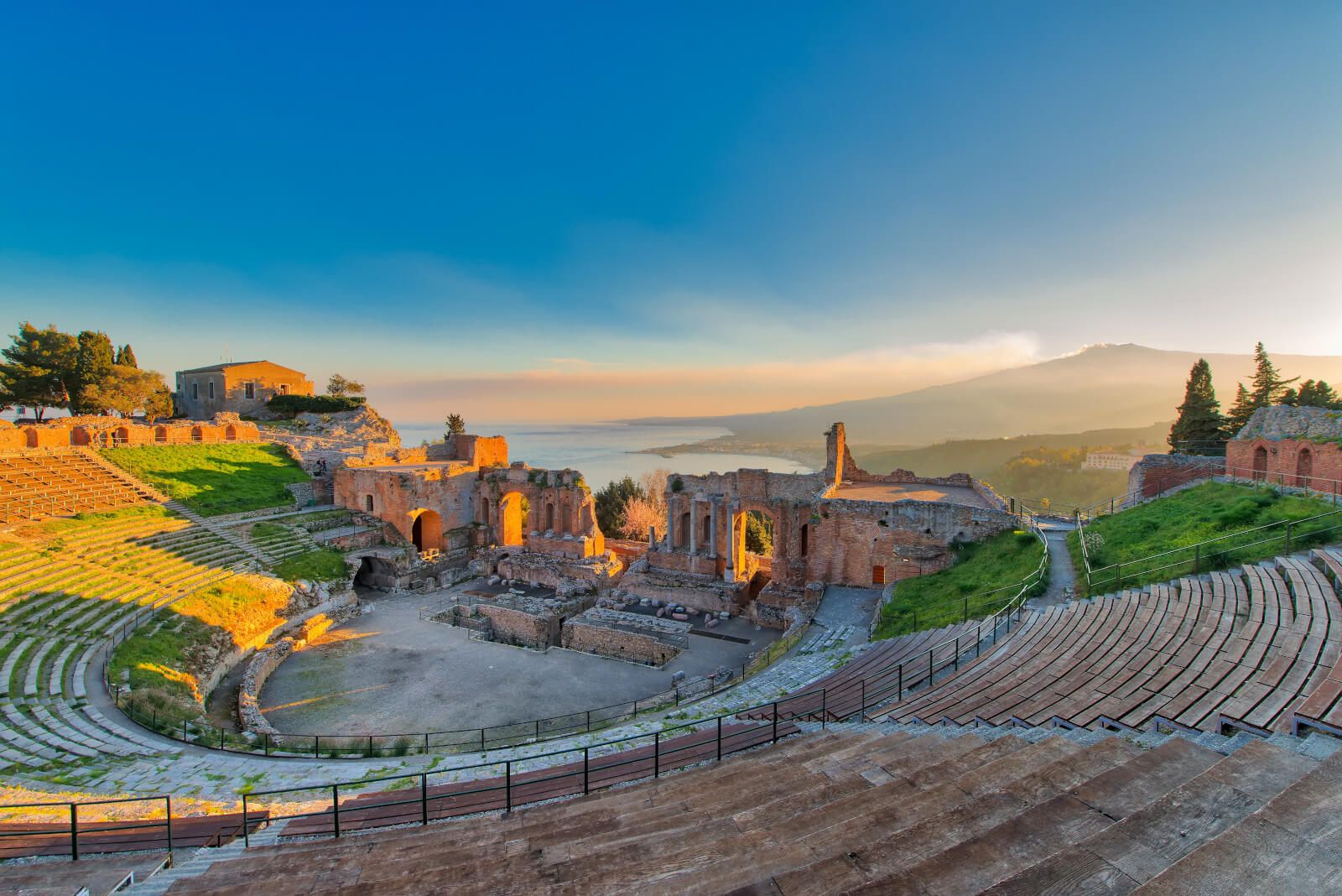 Ein antikes Theater mit Blick auf das Meer und den Etna im Hintergrund, умgeben von Ruinen und grünem Gras.