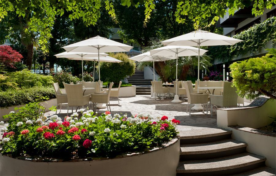 Gemütlicher Außenbereich mit Tischen, Stühlen und Sonnenschirmen, umgeben von bunten Blumen und üppigem Grün.
