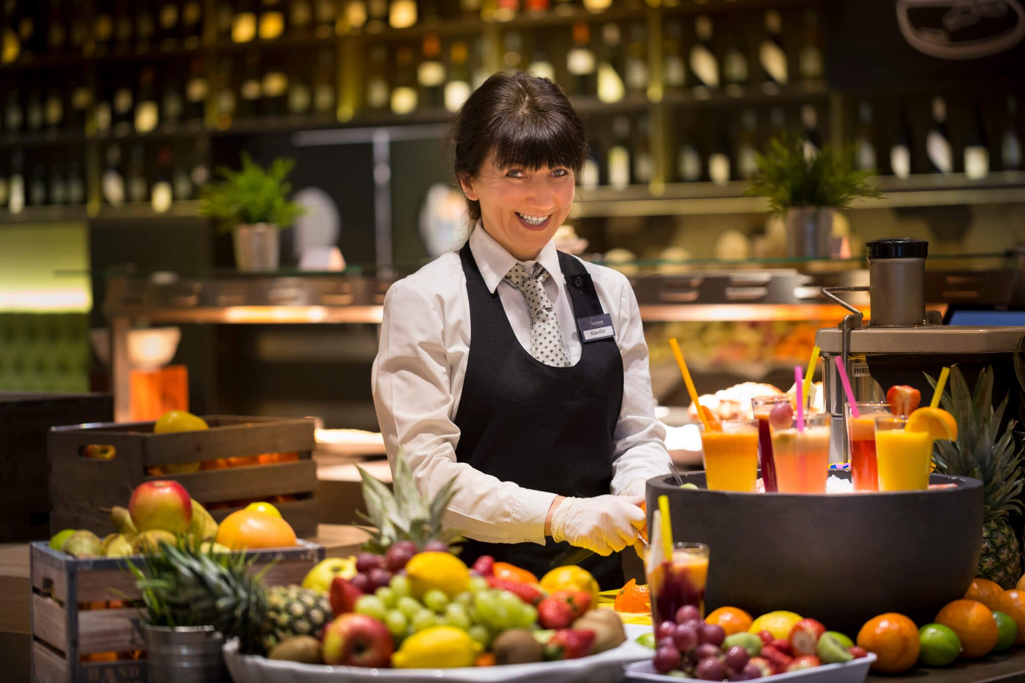 Eine fröhliche Bedienung steht an einem Tisch voller frischer Früchte und bunter Getränke in einem Restaurant.