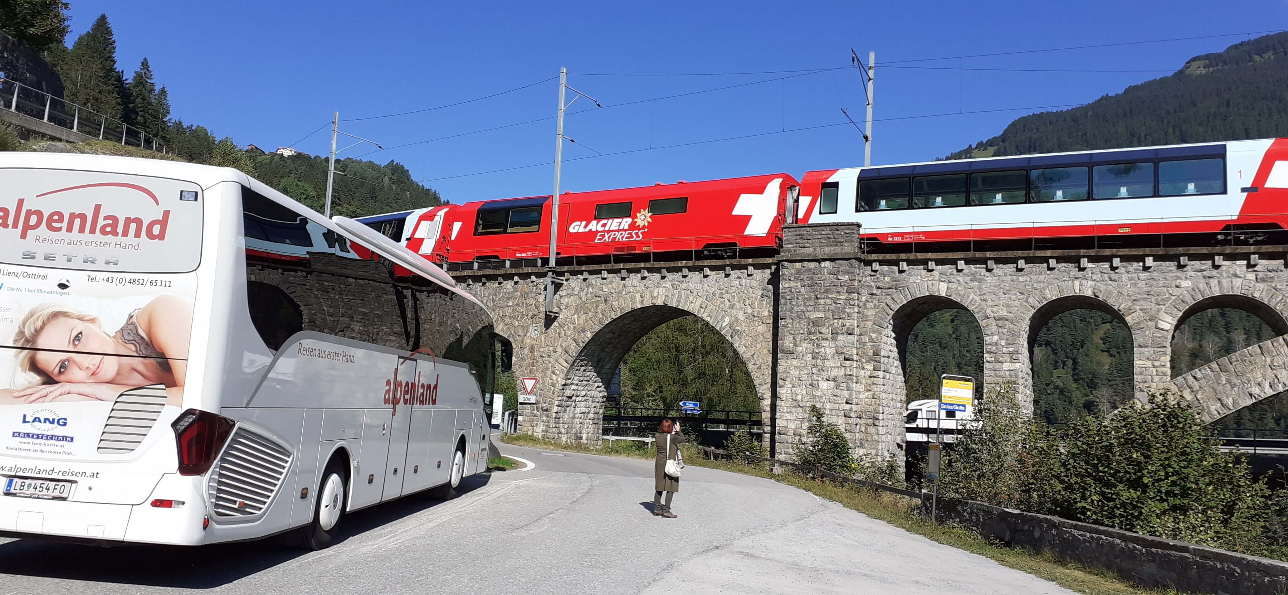 Ein Bus hält an einer Straße, während der Glacier Express auf einer Brücke darüber fuhr. Klare, sonnige Atmosphäre.