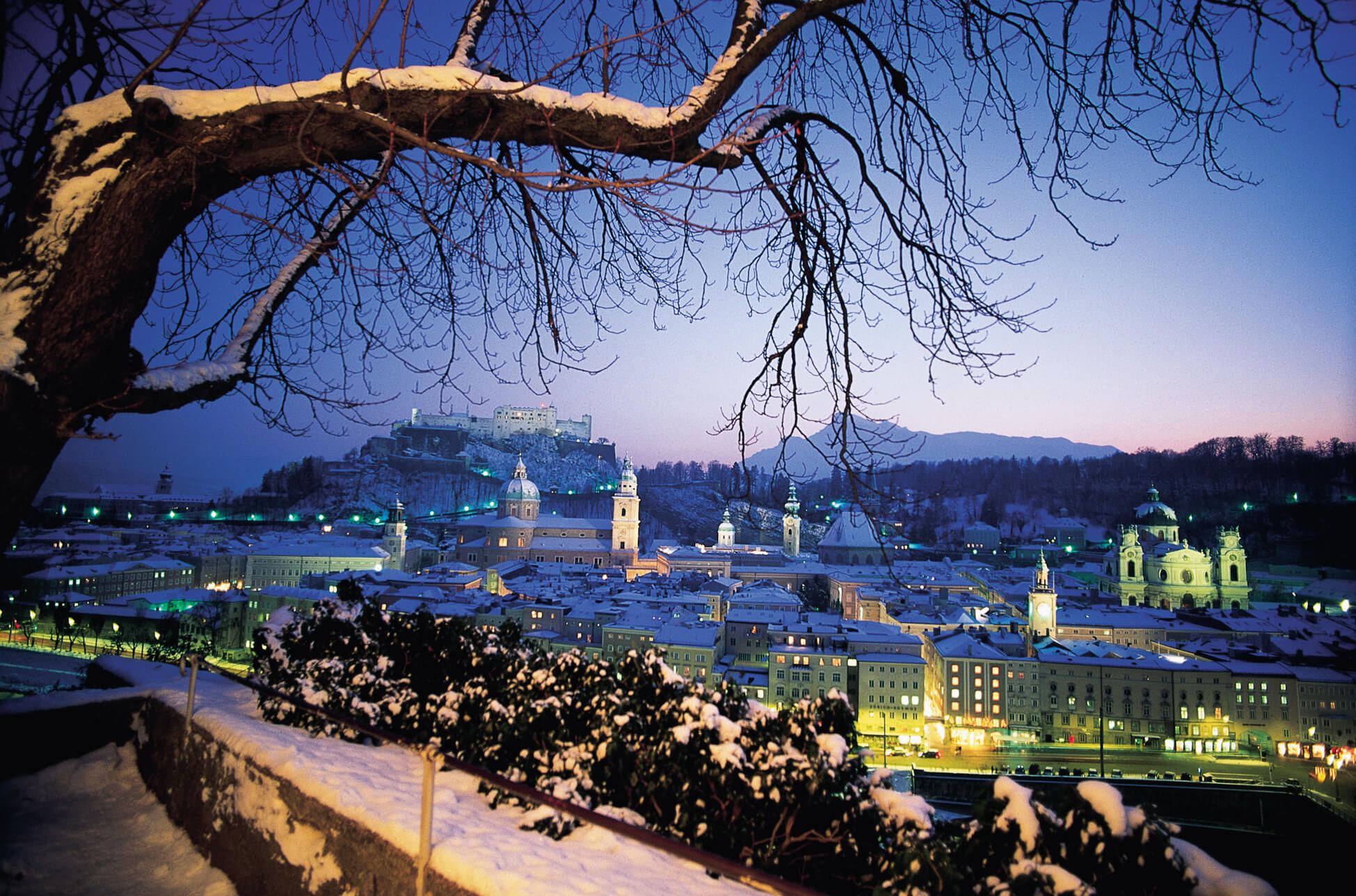Schneebedeckte Stadtansicht von Salzburg bei Dämmerung, mit einem Blick auf die Festung und glühenden Lichtern.