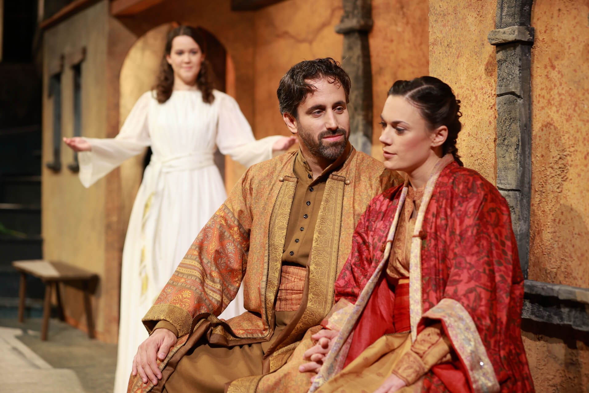 Drei Darsteller auf einer Bühne in historischen Kostümen, eine spricht, während die anderen aufmerksam lauschen.