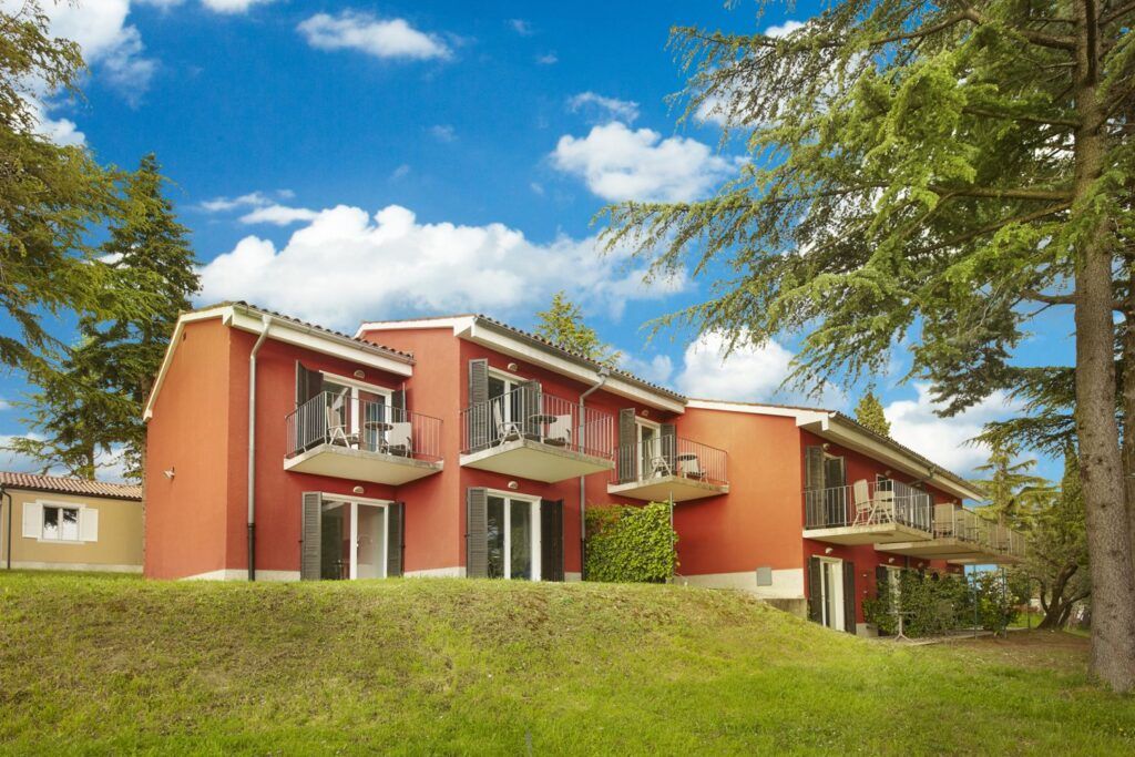 Zwei rote Mehrfamilienhäuser mit Balkonen, umgeben von Bäumen und blauem Himmel. Grüner Rasen im Vordergrund.