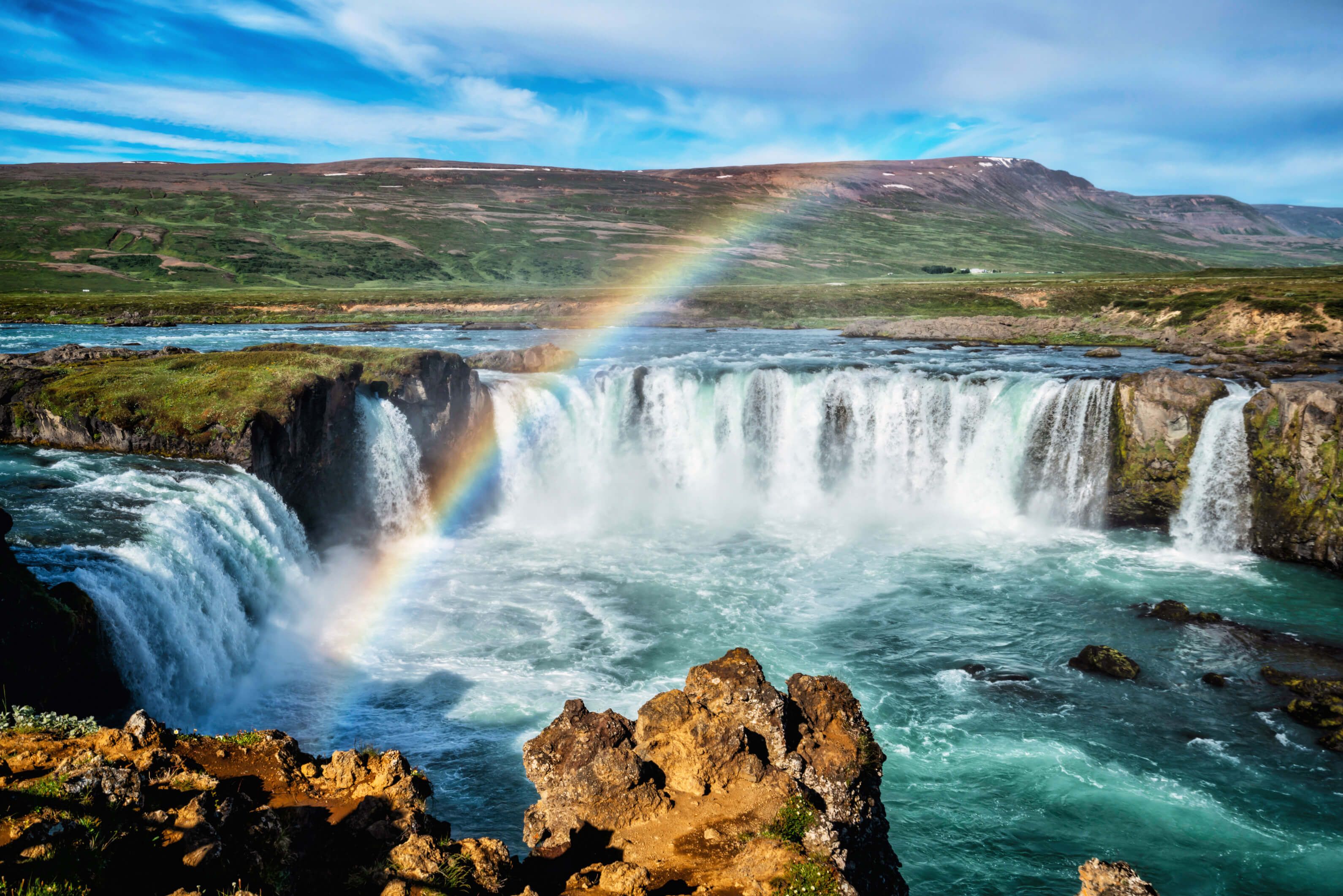 Ein wunderschöner Wasserfall mit Regenbogen und grüner Landschaft im Hintergrund. Klarer blauer Himmel darüber.