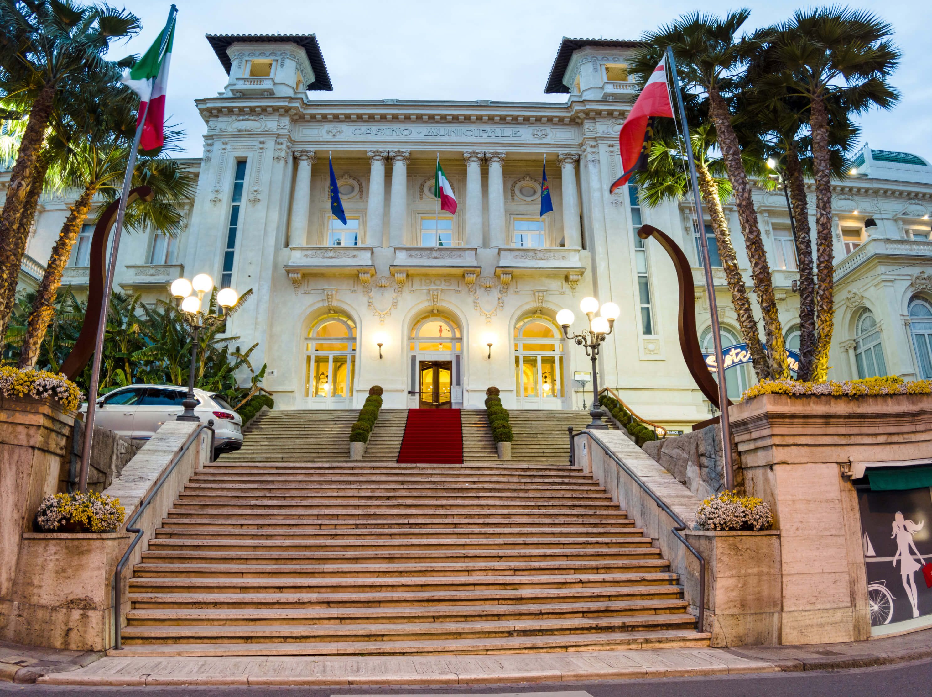 Ein prächtiges Casino mit Stufen, Palmen und Flaggen im abendlichen Licht. Schöne Architektur und gepflegte Umgebung.
