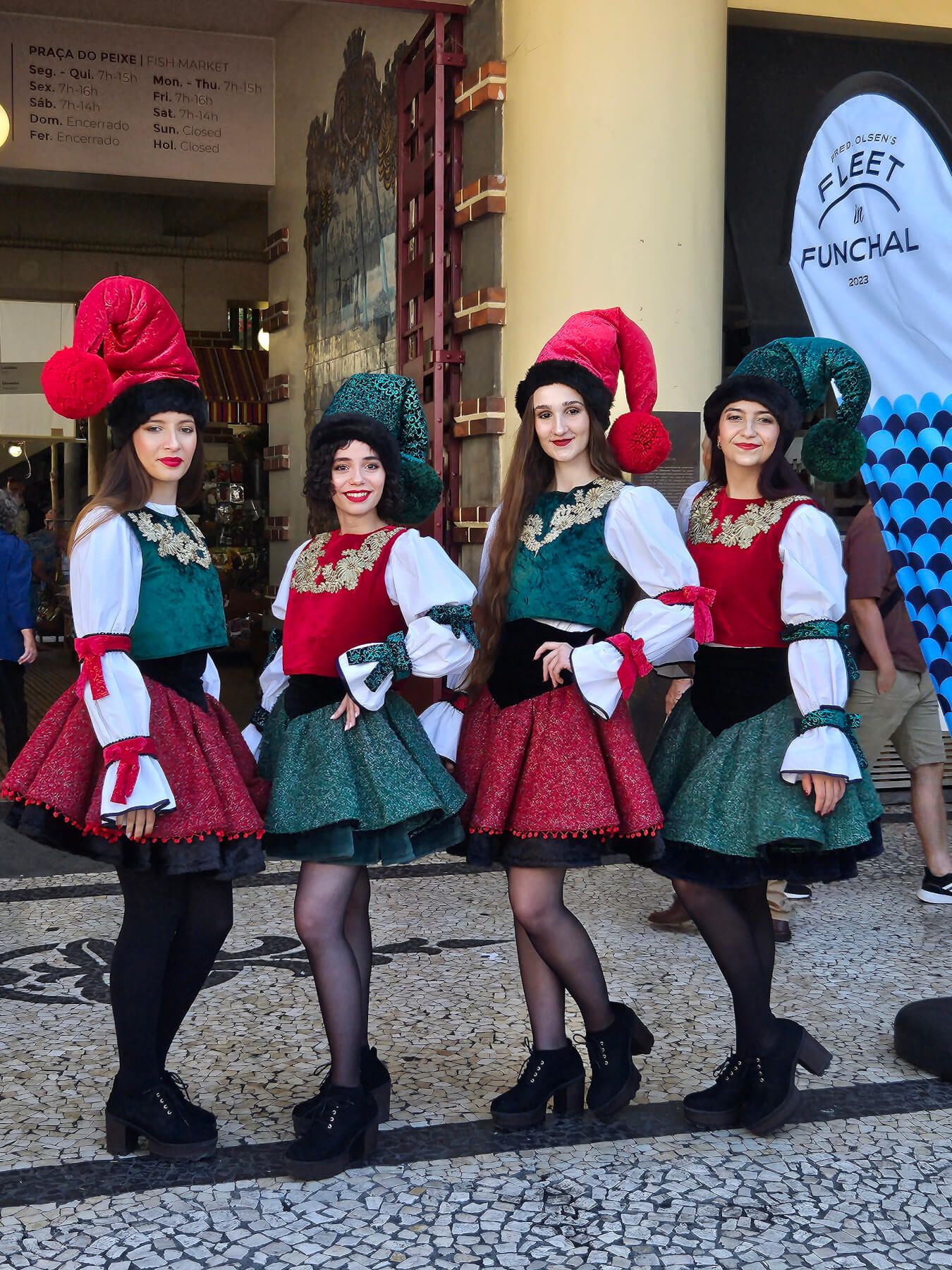 Vier Frauen in bunten traditionellen Kleidern und großen Hüten posieren fröhlich vor einem Markt.