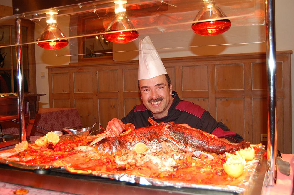 Ein Koch in einer weißen Mütze steht stolz neben einem großen, gebratenen Fisch auf einem Buffet.