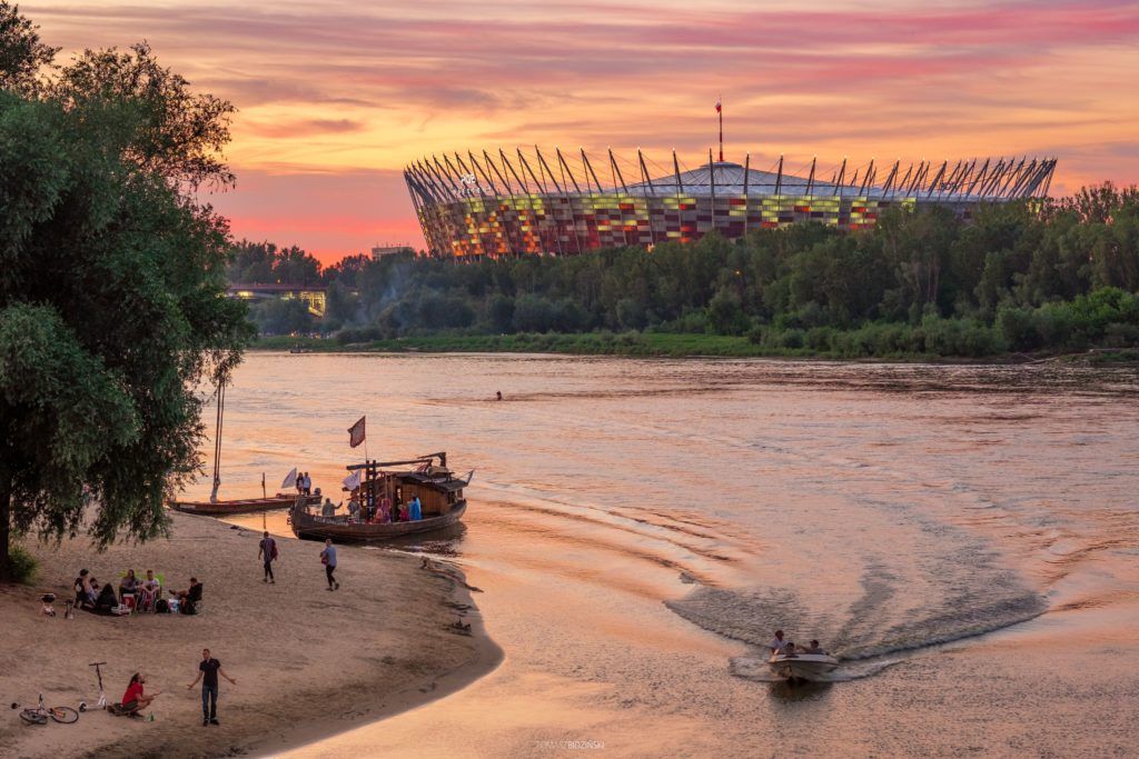 Ein farbenfroher Sonnenuntergang über einem Fluss mit einem Stadion im Hintergrund und Menschen am Ufer.