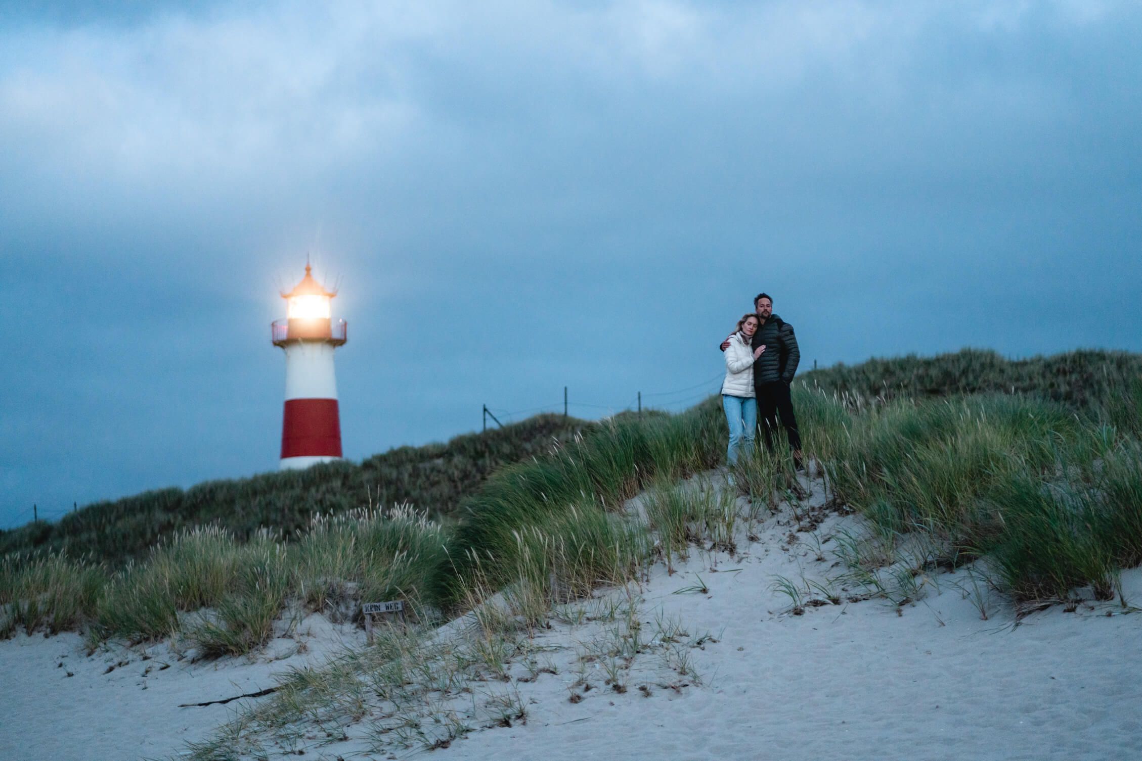 Ein Paar steht in den Dünen, der Leuchtturm leuchtet im Hintergrund bei dämmerigem Licht.