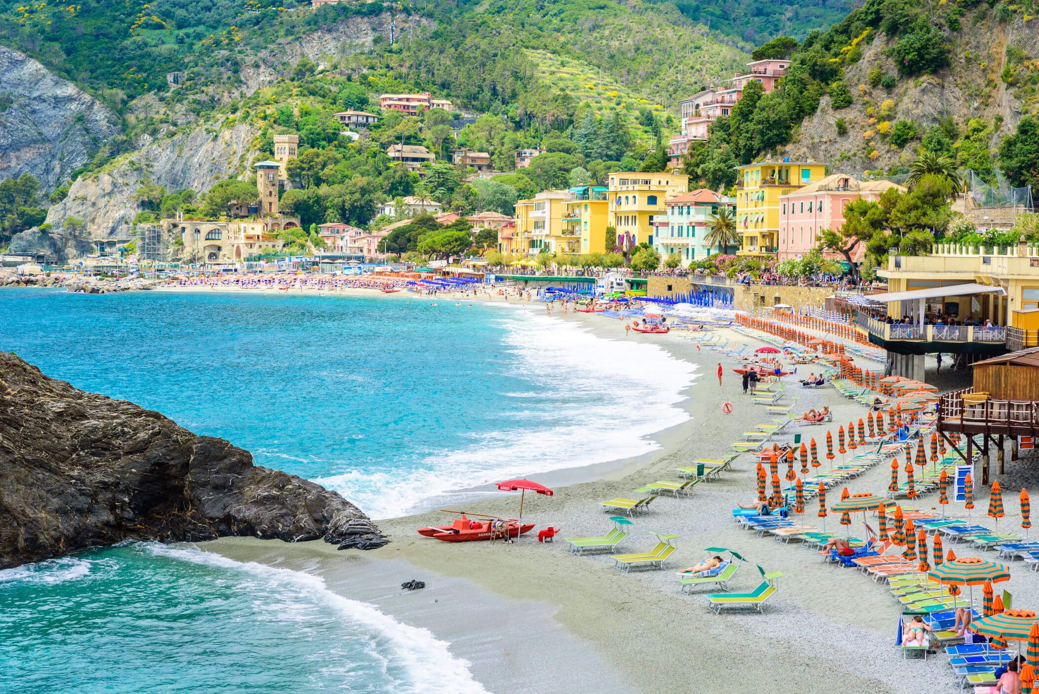 Ein sonniger Strand mit bunten Liegen, Sonnenschirmen und einem klaren blauen Meer. Hügel und farbige Gebäude im Hintergrund.