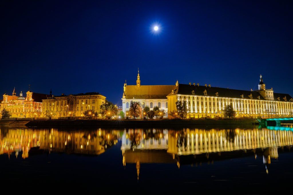 Eine nächtliche Stadtansicht mit beleuchteten Gebäuden und dem Mond, die sich im ruhigen Wasser widerspiegeln.