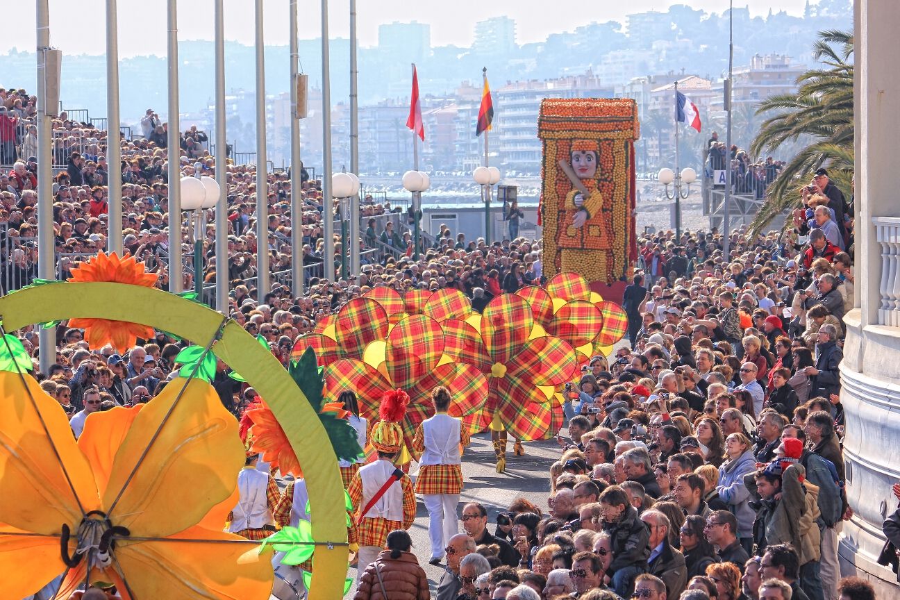 Eine bunte Parade mit vielen Menschen und festlichen Kostümen, umgeben von Blumen und einer auffälligen Figur.