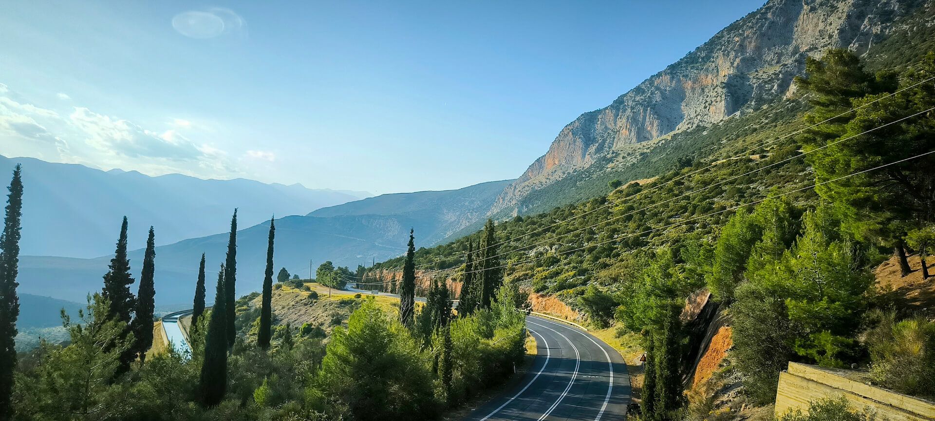Eine kurvenreiche Straße schlängelt sich durch bewaldete Hügel und Berge unter einem klaren blauen Himmel.